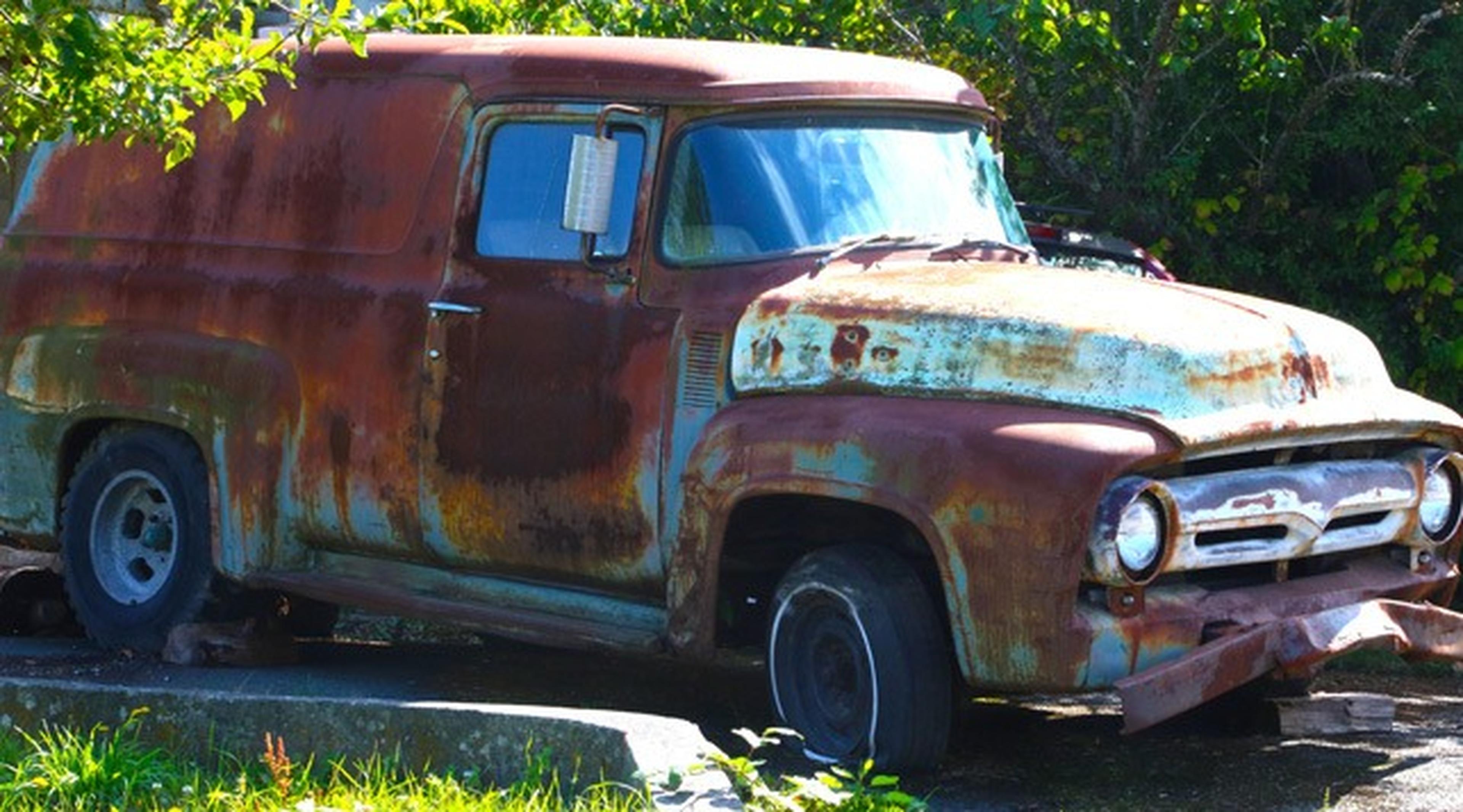 A rusty red truck sitting in a driveway surrounded by some green trees and grass