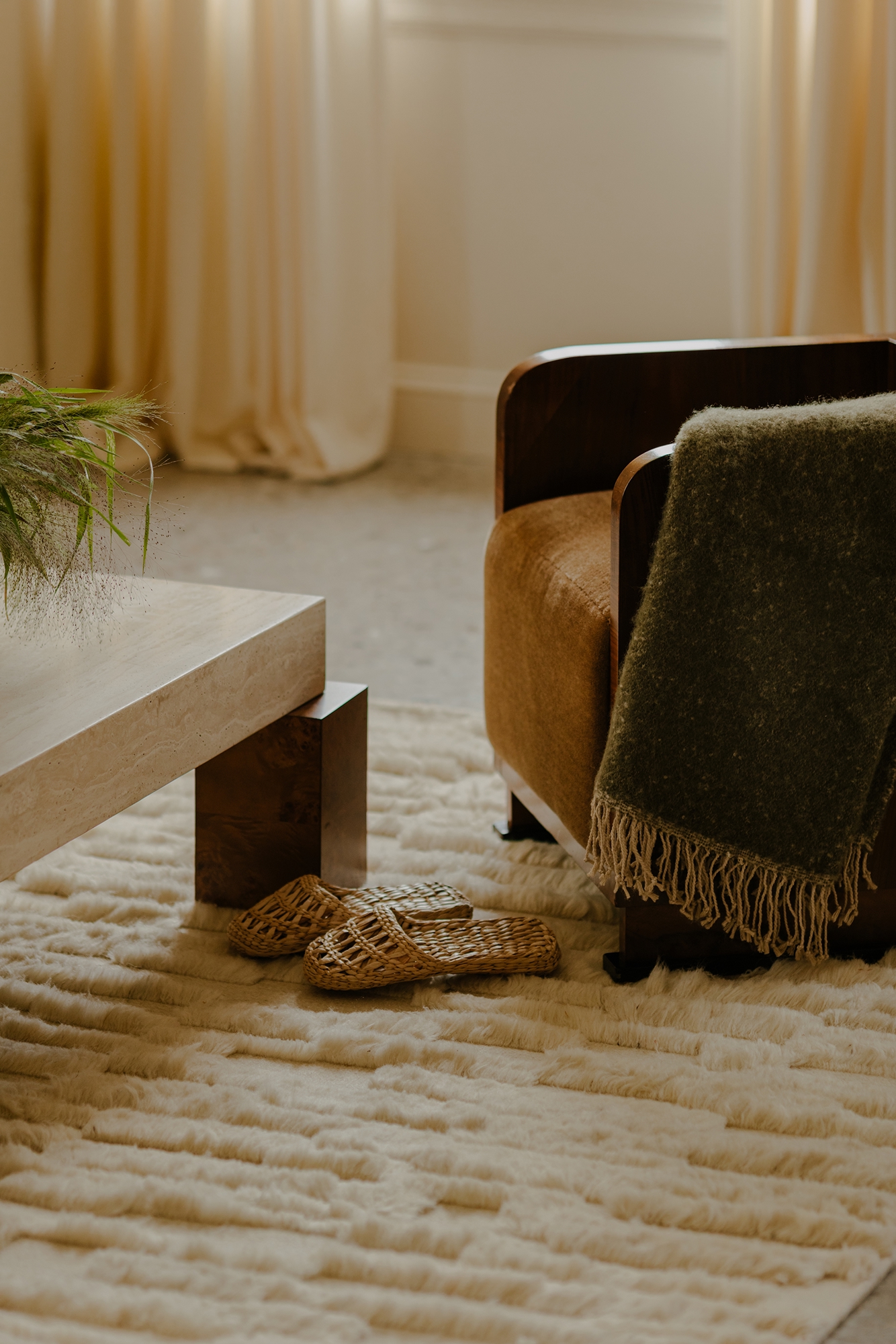 Cozy living room with a brown armchair draped in a green blanket, wicker slippers on a plush rug beside a wooden coffee table.