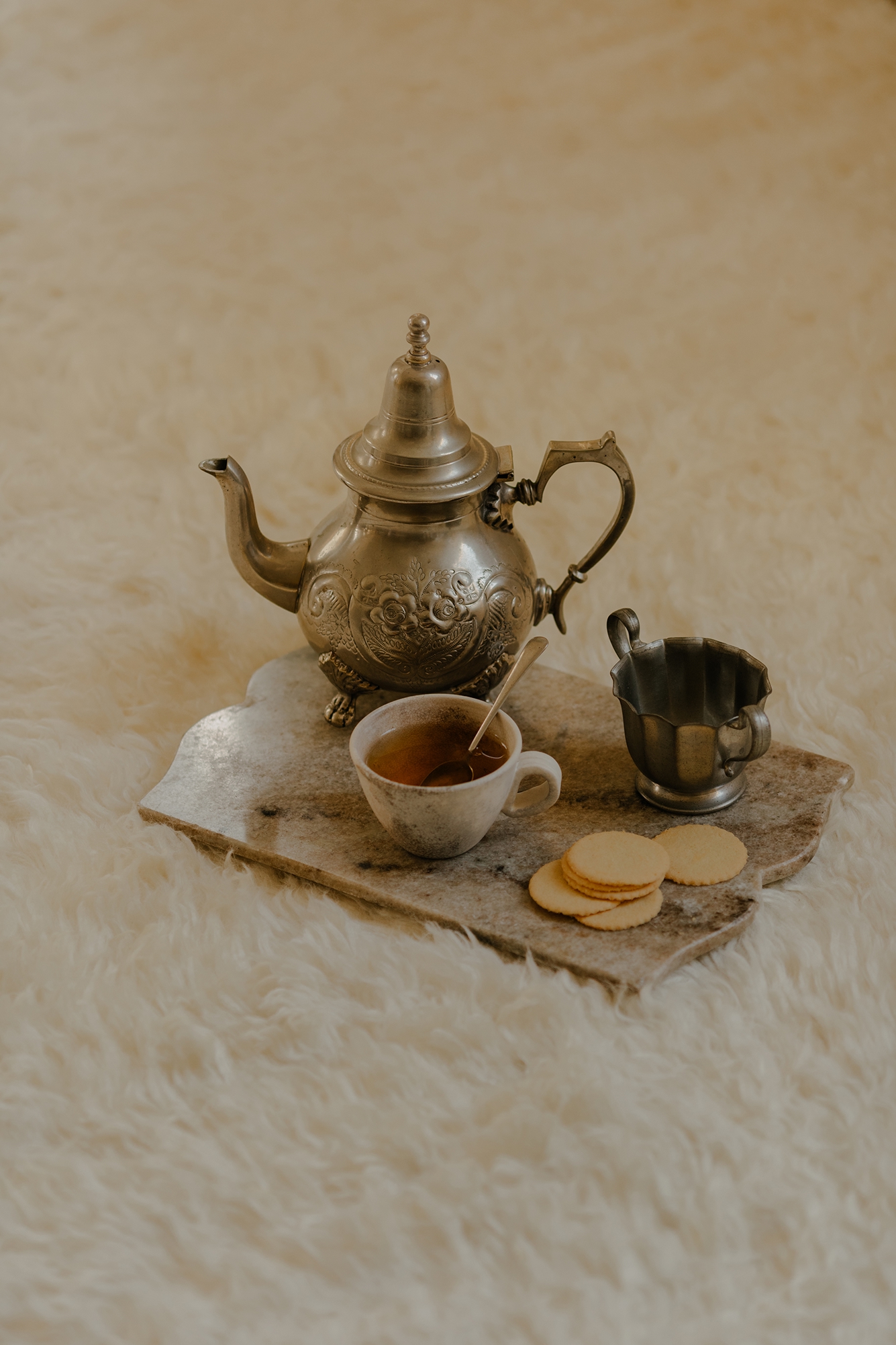 A silver teapot, a mug with tea and a spoon, a small creamer, and five cookies on a stone tray resting on a fluffy white rug.