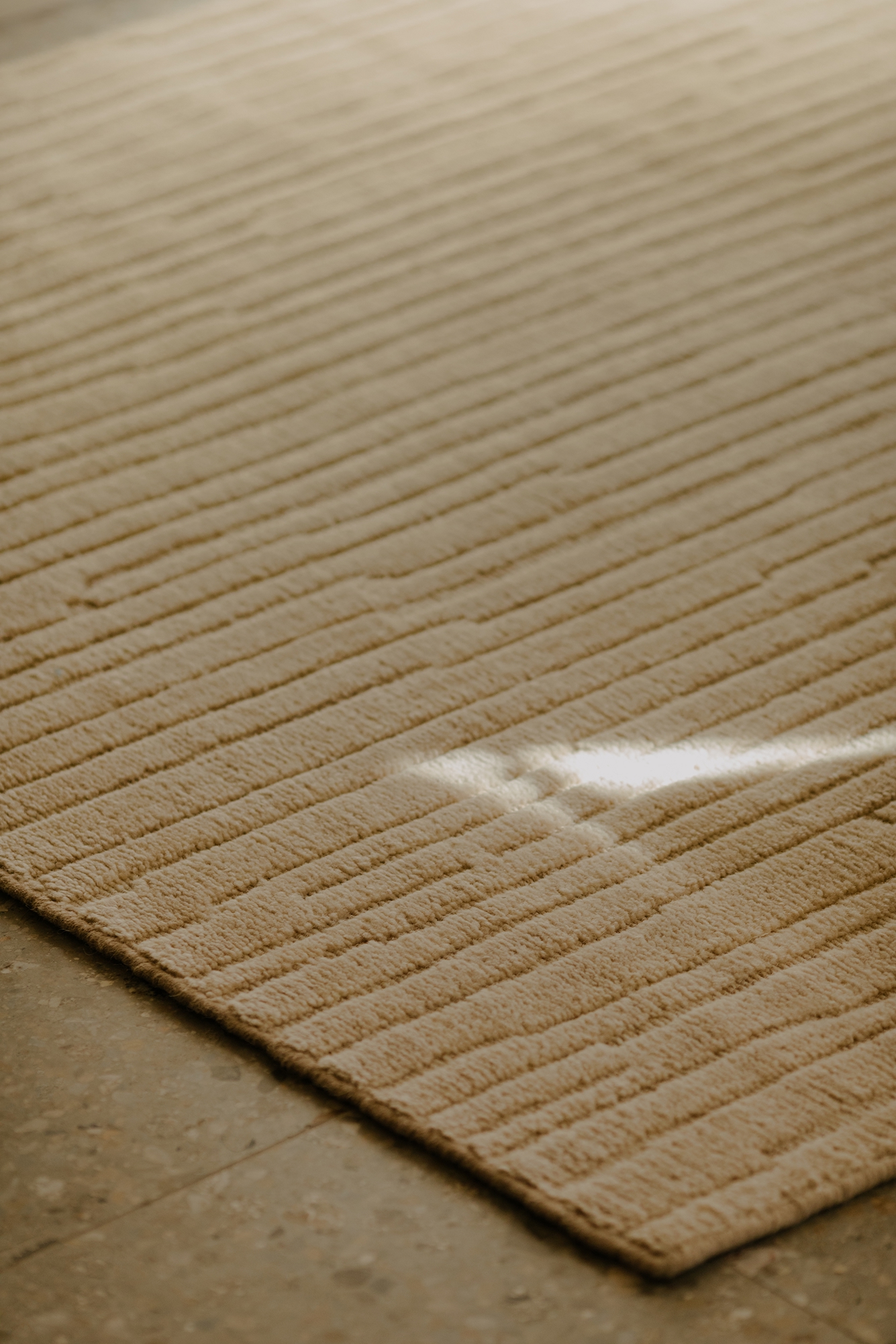 A beige textured rug with parallel lines on a stone floor, illuminated by a patch of sunlight.