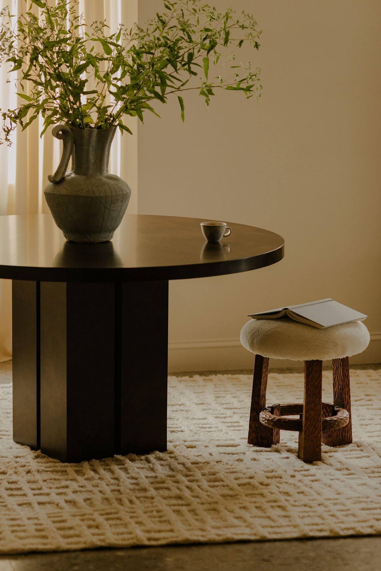A dark round table with a vase of greenery and a cup, next to a small stool with a book, on a textured ivory rug in a softly lit room.