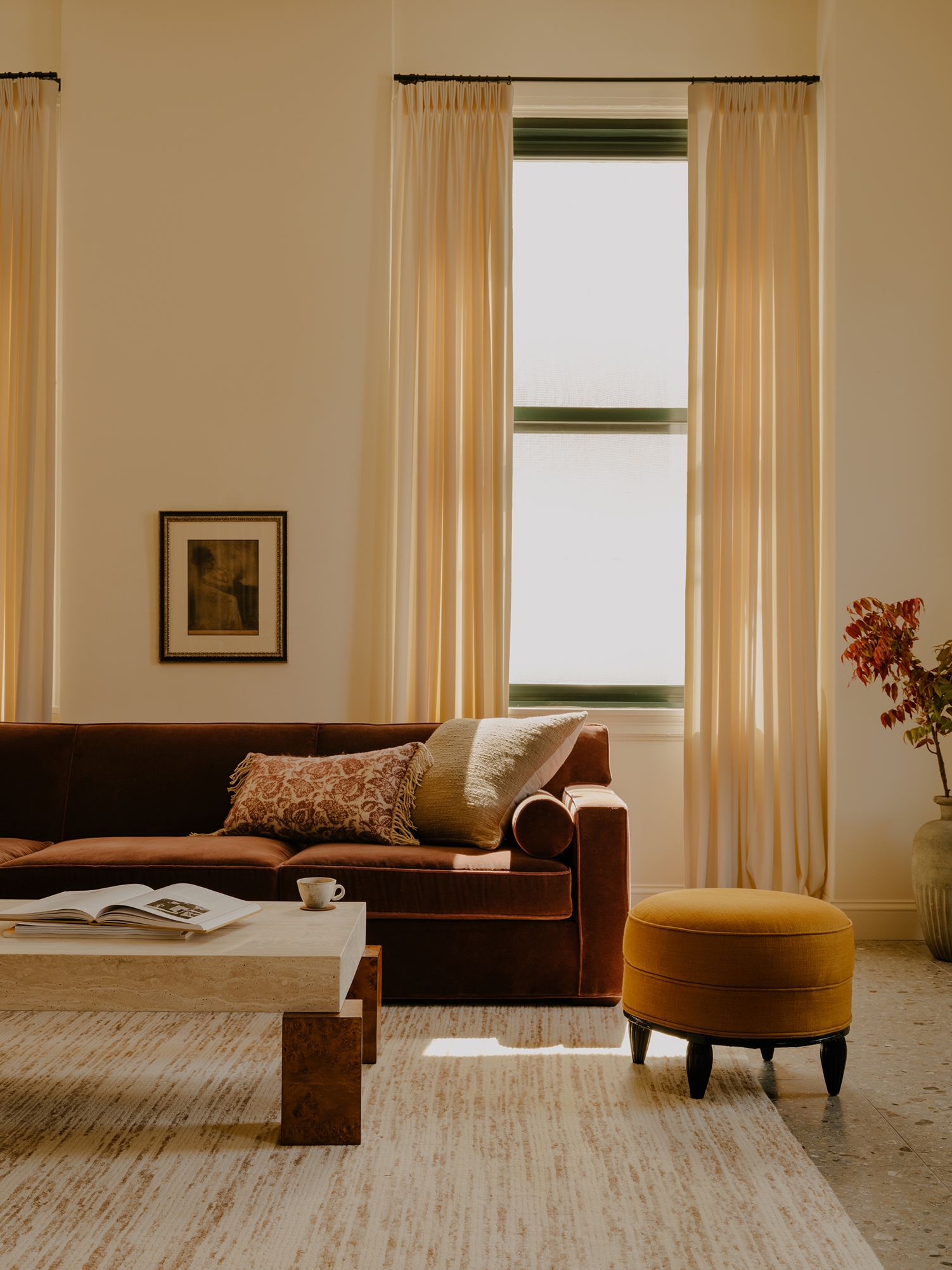 A cozy living room with a brown velvet sofa, a marble coffee table, and a mustard ottoman. Sunlight filters through tall cream curtains.