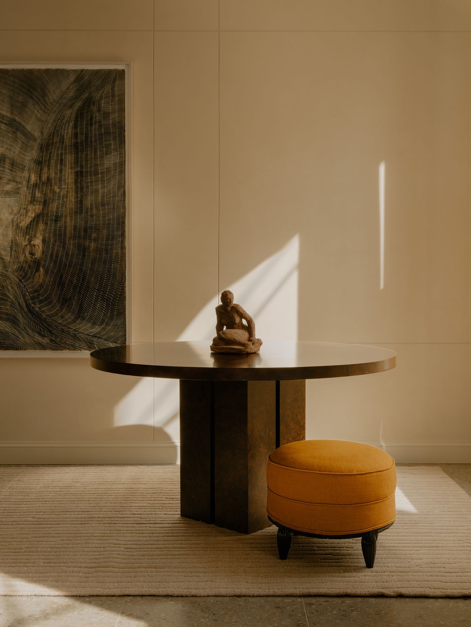A round table with a small sculpture, next to an orange ottoman, in a warmly lit room with abstract wall art, on an ivory rug with texture.