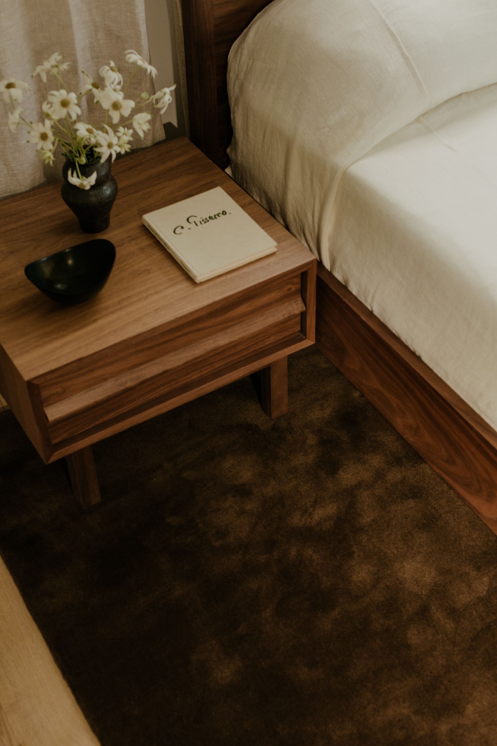 Dark brown rug over hardwood floors with a small side table and white bed over it 