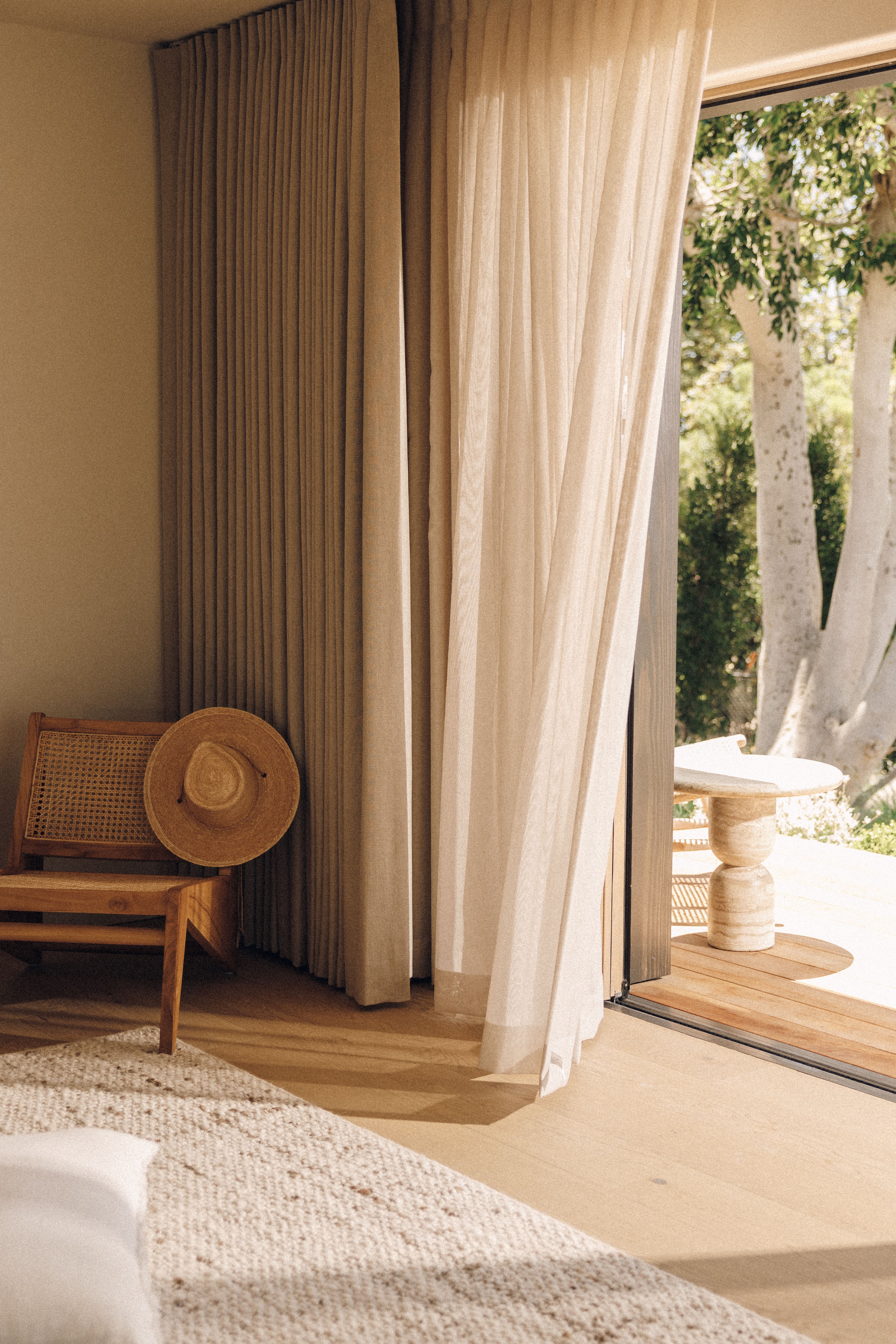 Cane chair with a straw sun hat beside linen curtains and open doors to a sunny deck, with Cormac in Oatmeal in the foreground