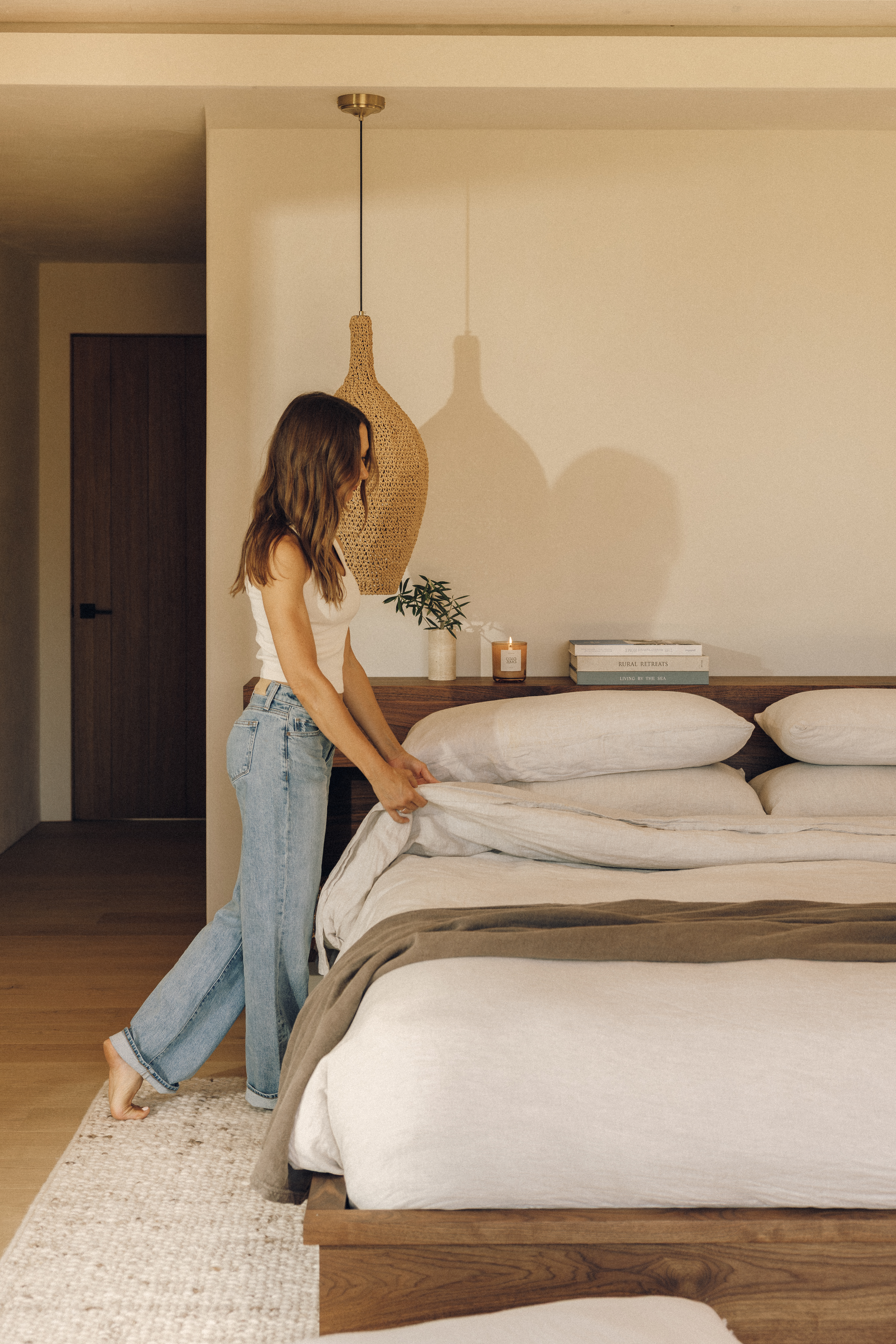 Woman smoothing white linen bedding on a low walnut bed beneath a woven pendant light, with Cormac in Oatmeal underfoot
