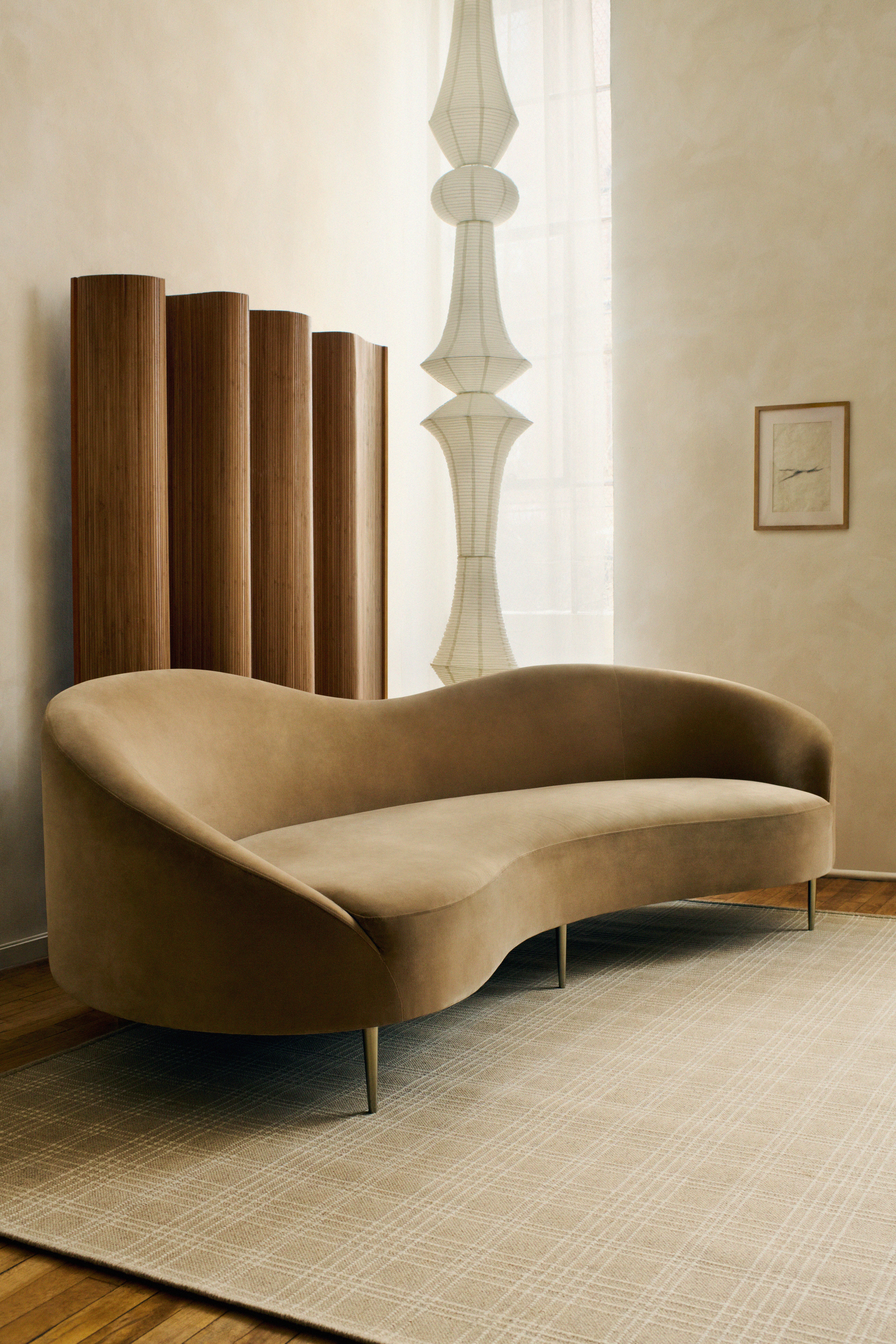 Elegant tan curved sofa in a minimalist room with a tall white lamp, wooden paneling, and framed artwork. Beige -- Ellis in Sand-- rug on wooden floor.