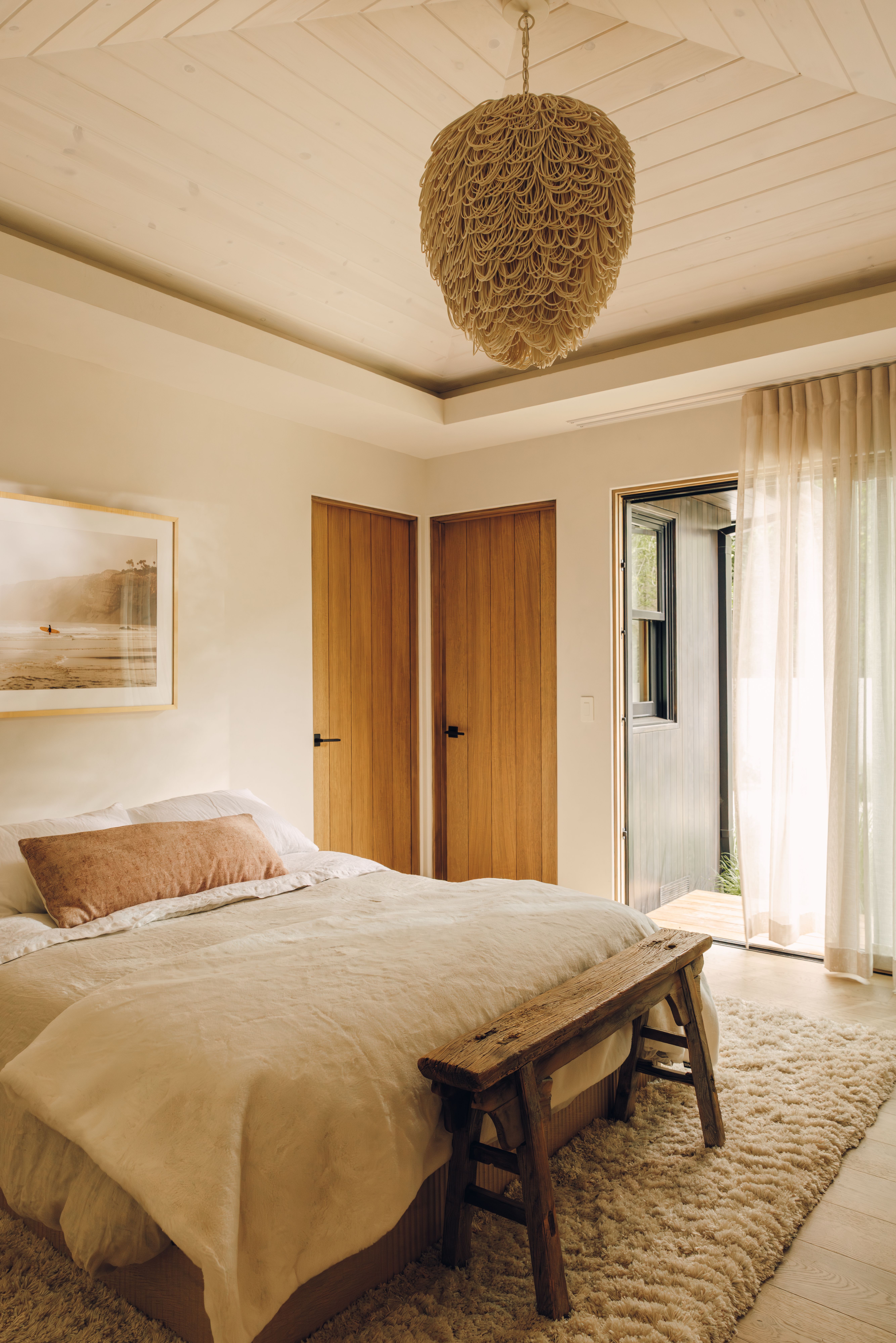 Bedroom with an oversized woven pendant, cream linen bedding and a rustic wood bench, with Sugar in Cream underfoot