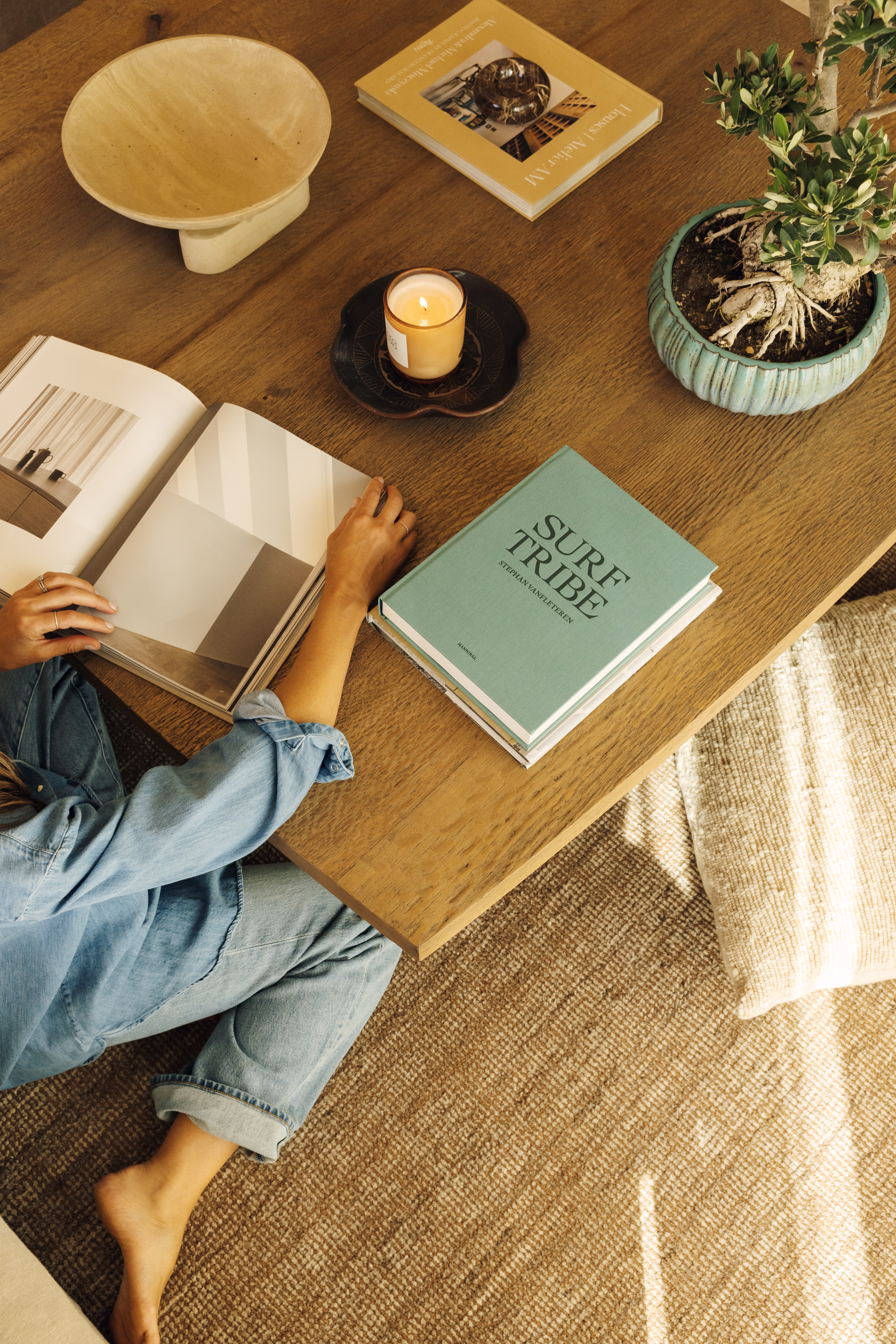 Person in denim reading on the floor at a wood coffee table with books, a candle and a potted plant, over Xander in Cedar
