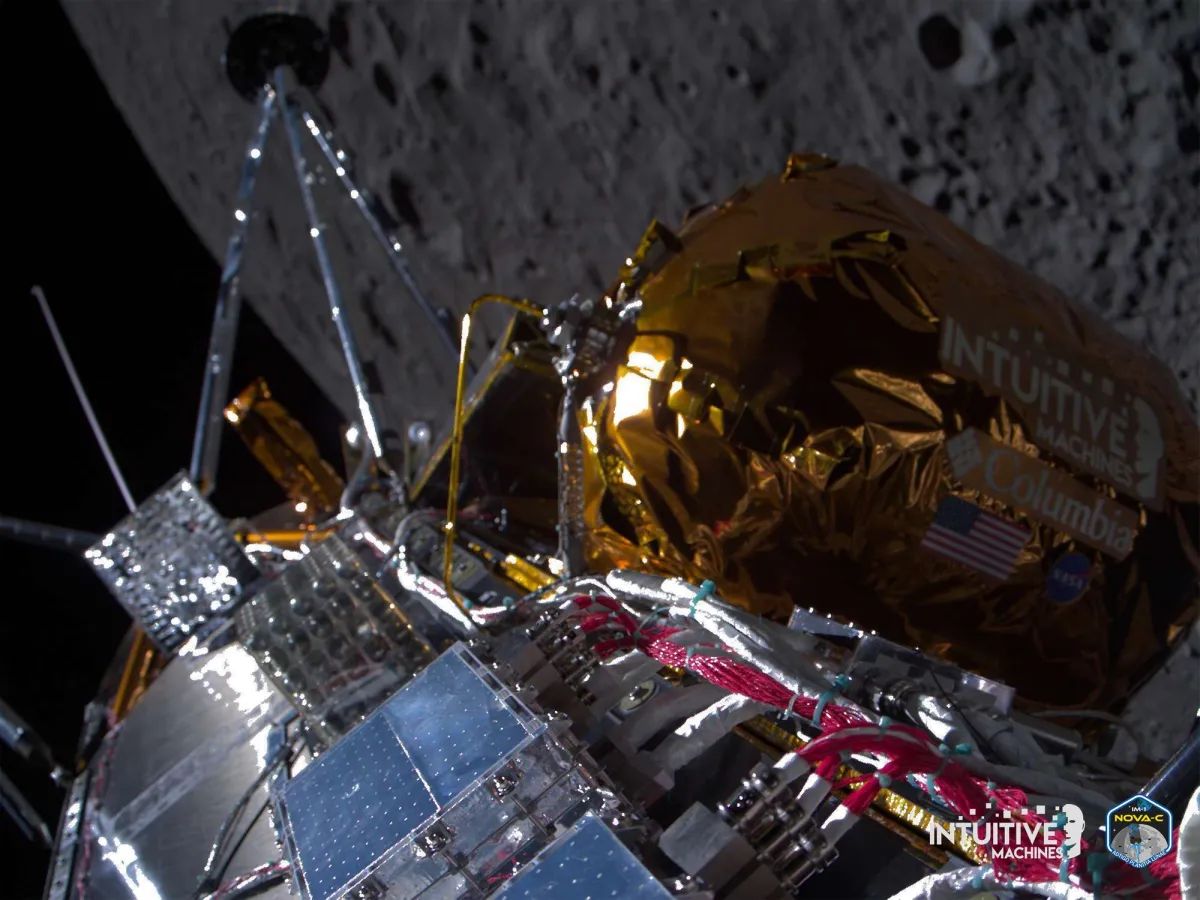 A camera view looking down on a robotic spacecraft, orbiting over the Moon. Shiny gold and metallic parts are visible around solar panels and landing legs, as well as images showing the logos of Intuitive Machines, Columbia, NASA and a United States Flag. In the background, the crater-marked surface of the Moon is visible.