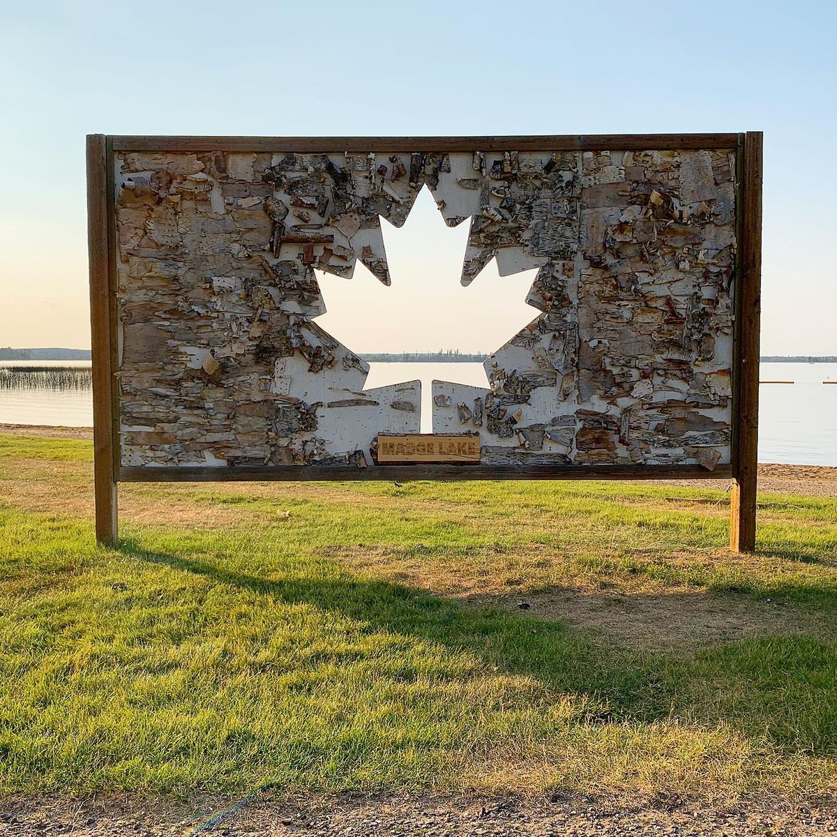 A wooden sign made out of scraps of wood and tree bark next to a lake in Saskatchewant. A maple leaf is carved out of the middle of the sign, allowing light to pass through. The placard beneath it says Madge Lake,