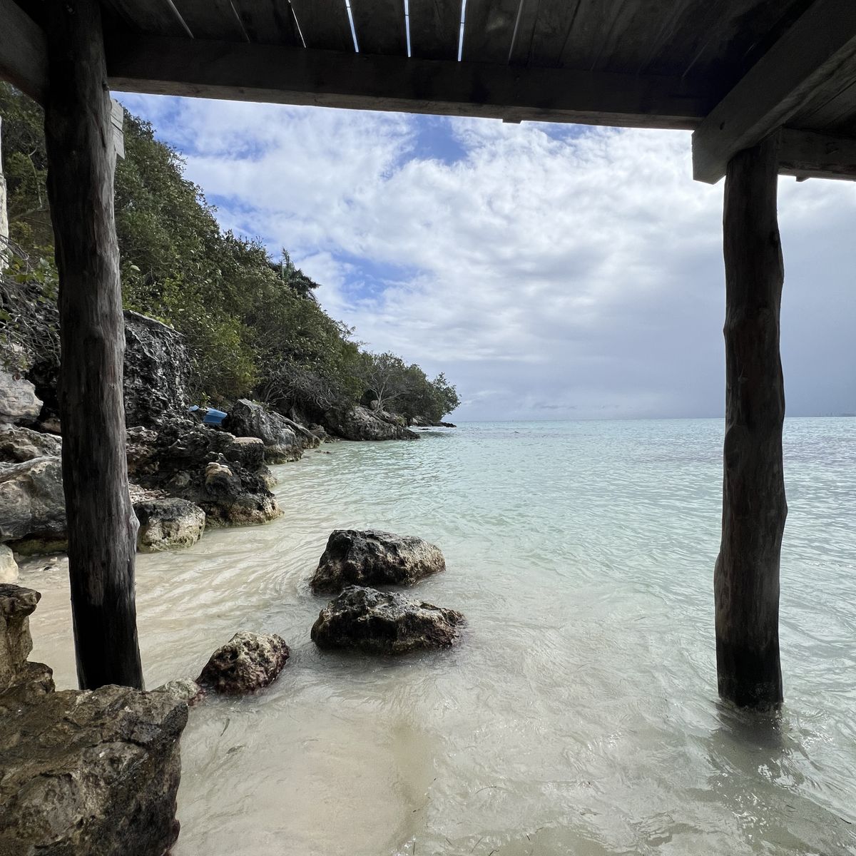 A rocky beach in southern Mexico, with a tree covered hill overlooking it. The phot is taken from underneath a wooden pier.