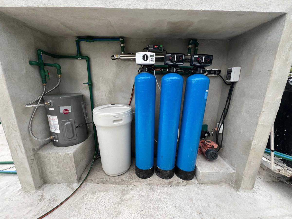 A concrete shed with a full water filtration system, including a pump, three tall blue cylinders, a grey garbage bin, a dark grey hot water heater, a long horizontal UV light and green plumbing joining everything together.