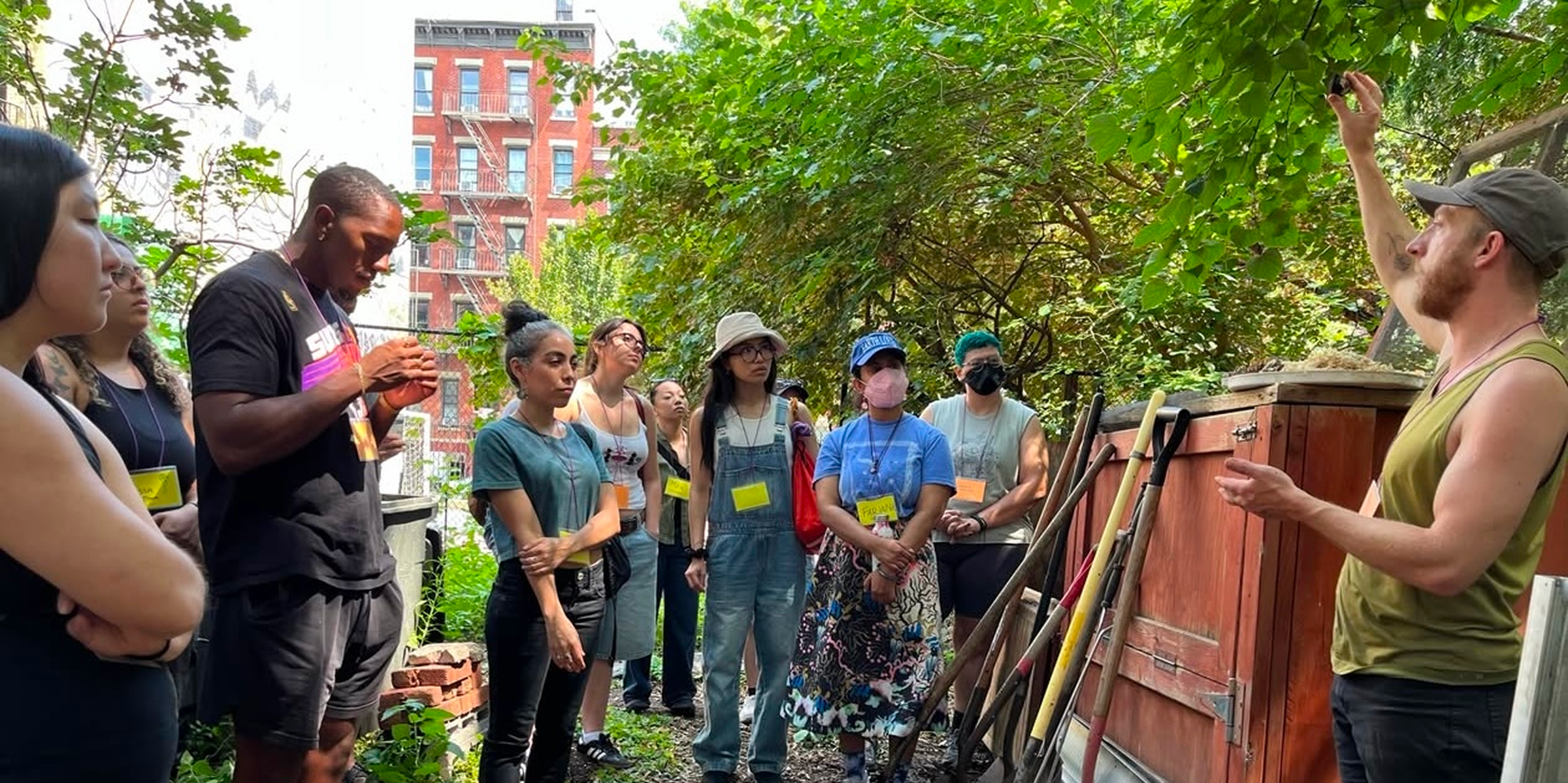 A group of people gathered outdoors, listening to a person speaking. They are surrounded by greenery next to garden tools and a building in the background.