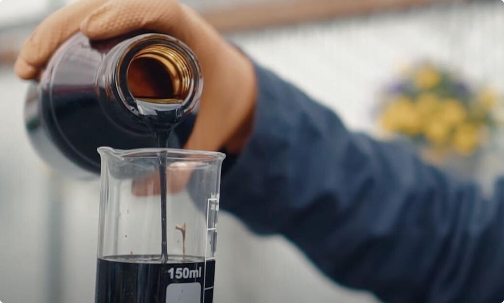 Person pouring thick, dark liquid from a brown bottle into a 150ml beaker, wearing a glove, with blurred background.