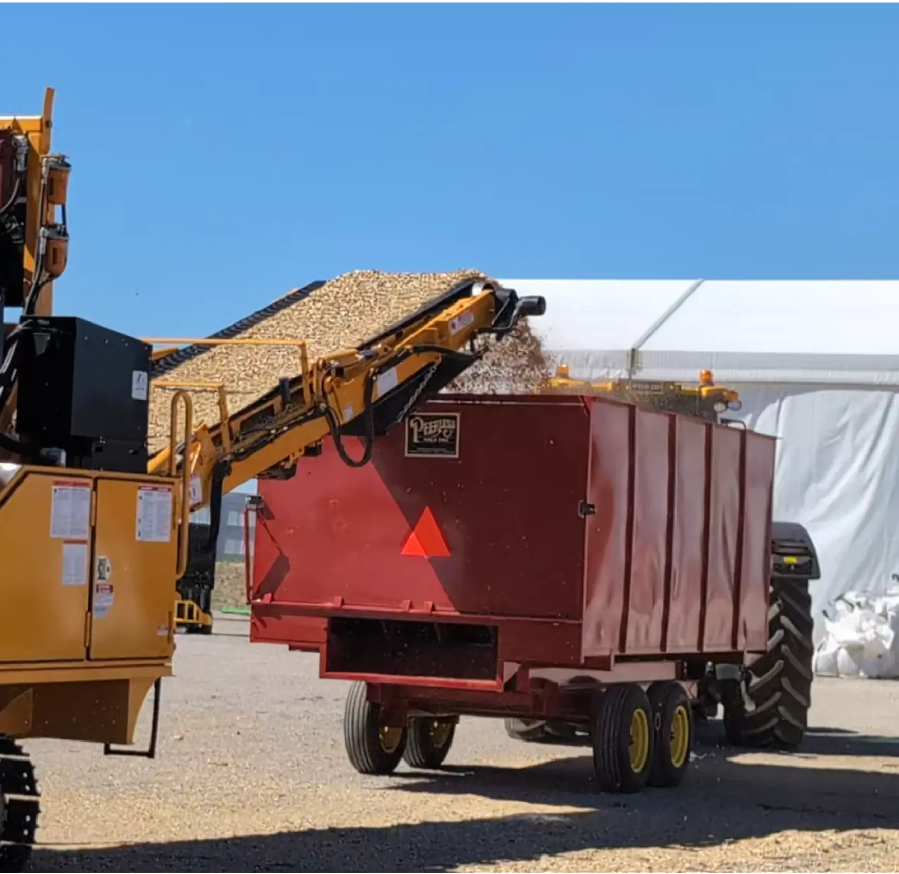 A machine unloads wood chips into a red trailer attached to a tractor on a sunny day, with a large tent in the background.