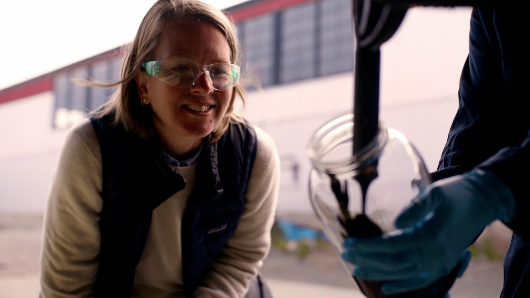 Jen, a Carbon Removal Geologist for Charm Industrial, smiles as she observes a sample of dark, viscous bio-oil being poured into a clear jar, highlighting the company's sequestration process.