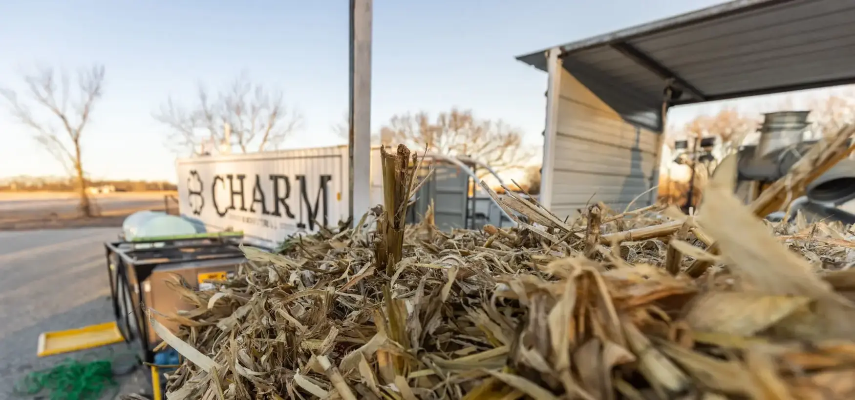 Close-up of dried corn stalks with a CHARM container and farm equipment in the background under a clear sky.
