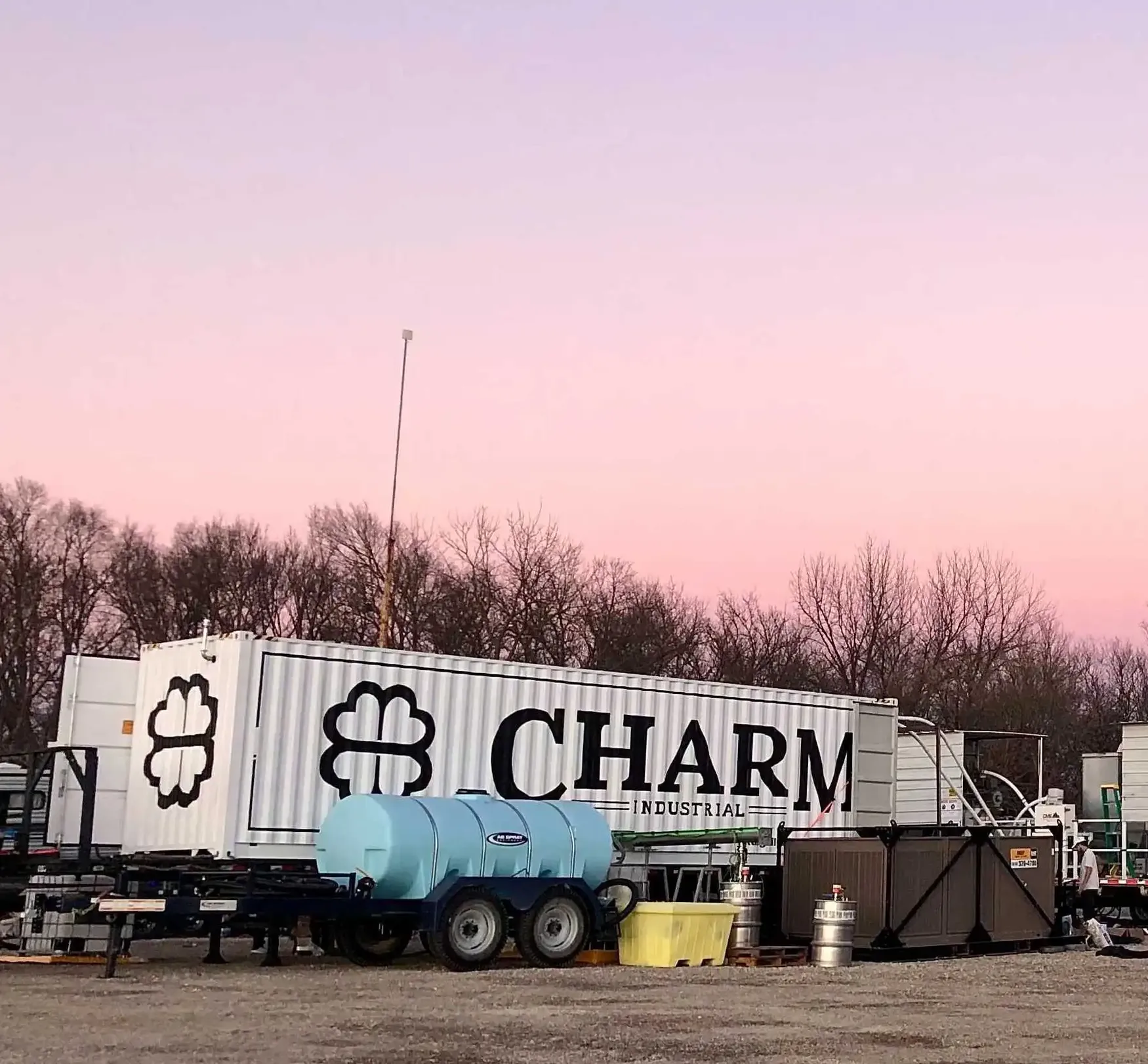 A Charm Industrial container and equipment are set up outdoors at sunset, with a pink sky and bare trees in the background.