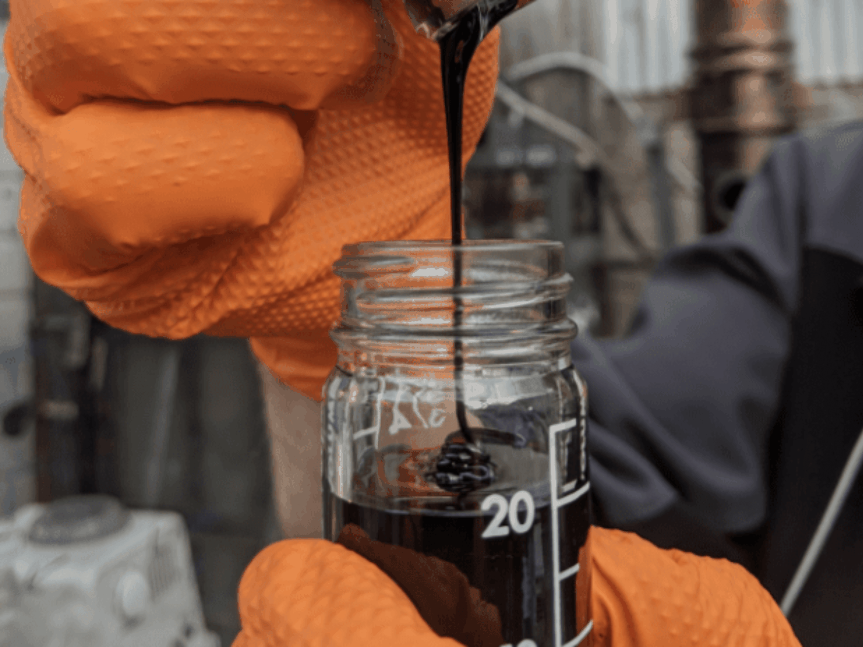 Person in orange gloves pouring thick black liquid into a glass jar with measurement markings.
