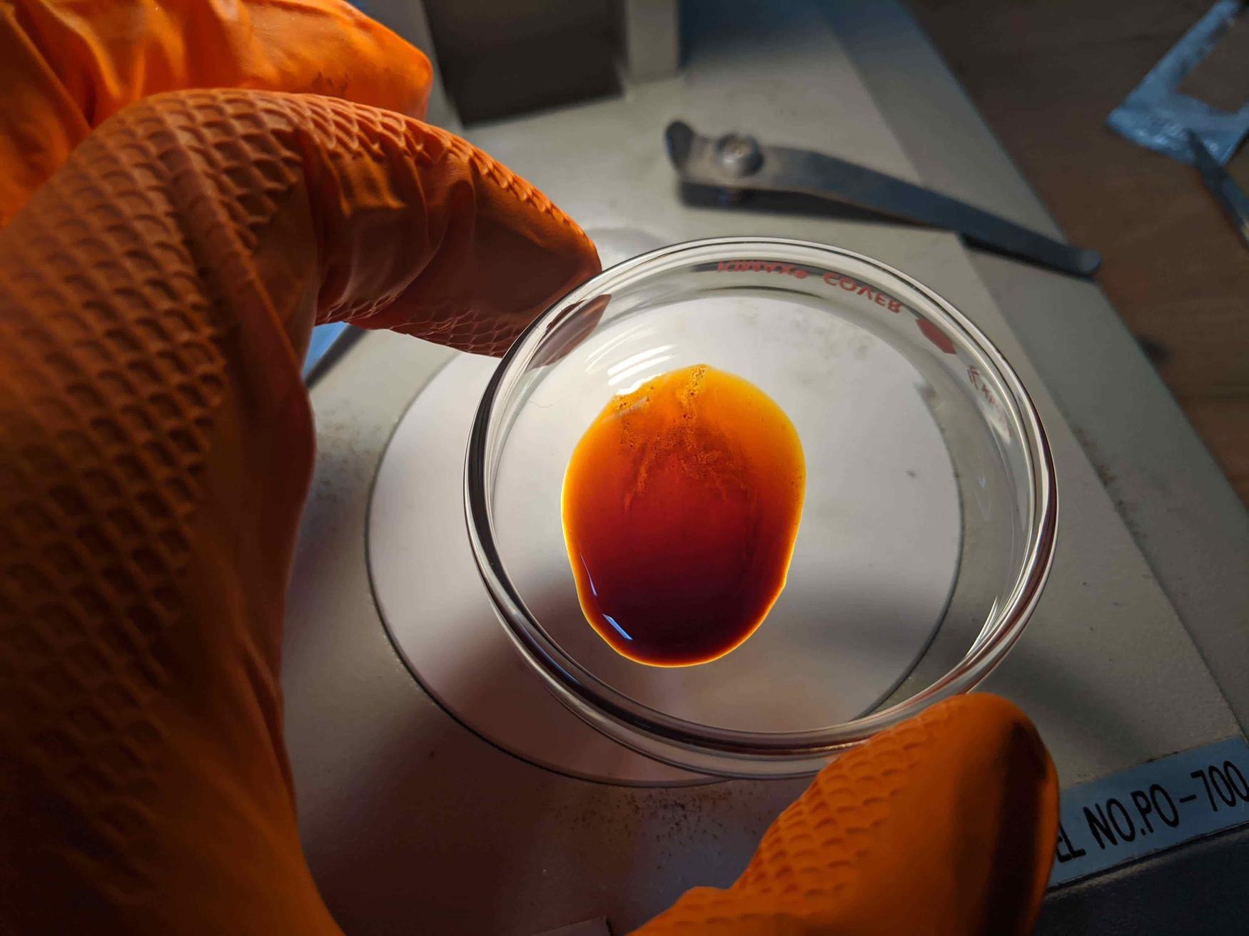 A person in orange gloves holds a small glass dish containing a reddish-brown liquid under a light, with a wrench in the background.