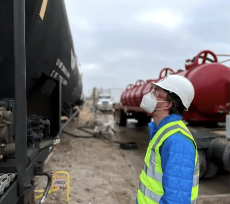 Worker in a hard hat and safety vest inspects a black train car, with red equipment in the background on a cloudy day.