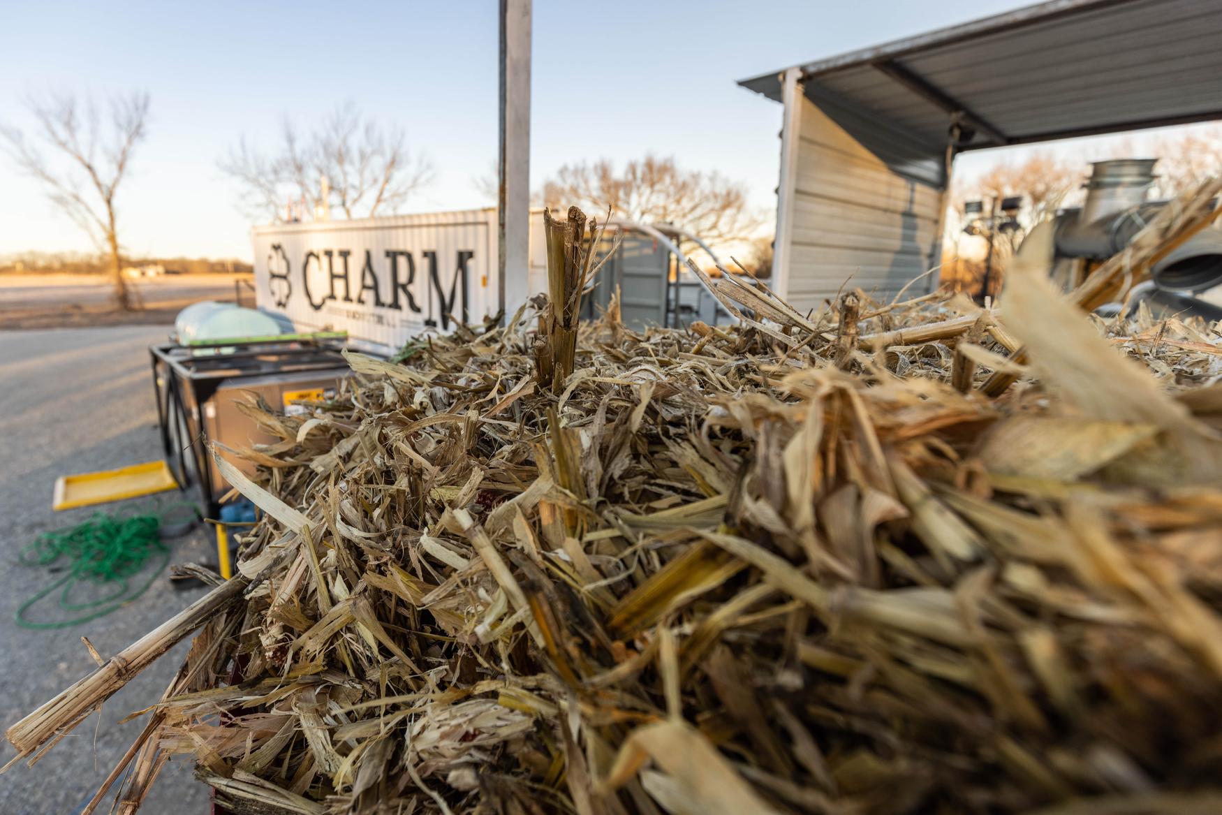 Close-up of a pile of dried corn husks with a shipping container labeled "CHARM" in the background, set in an outdoor industrial area.