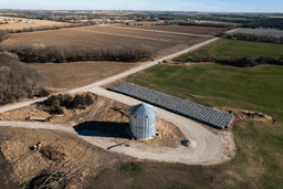 Aerial view of a rural landscape with a grain silo, solar panels, fields, and a winding dirt road under a clear blue sky.