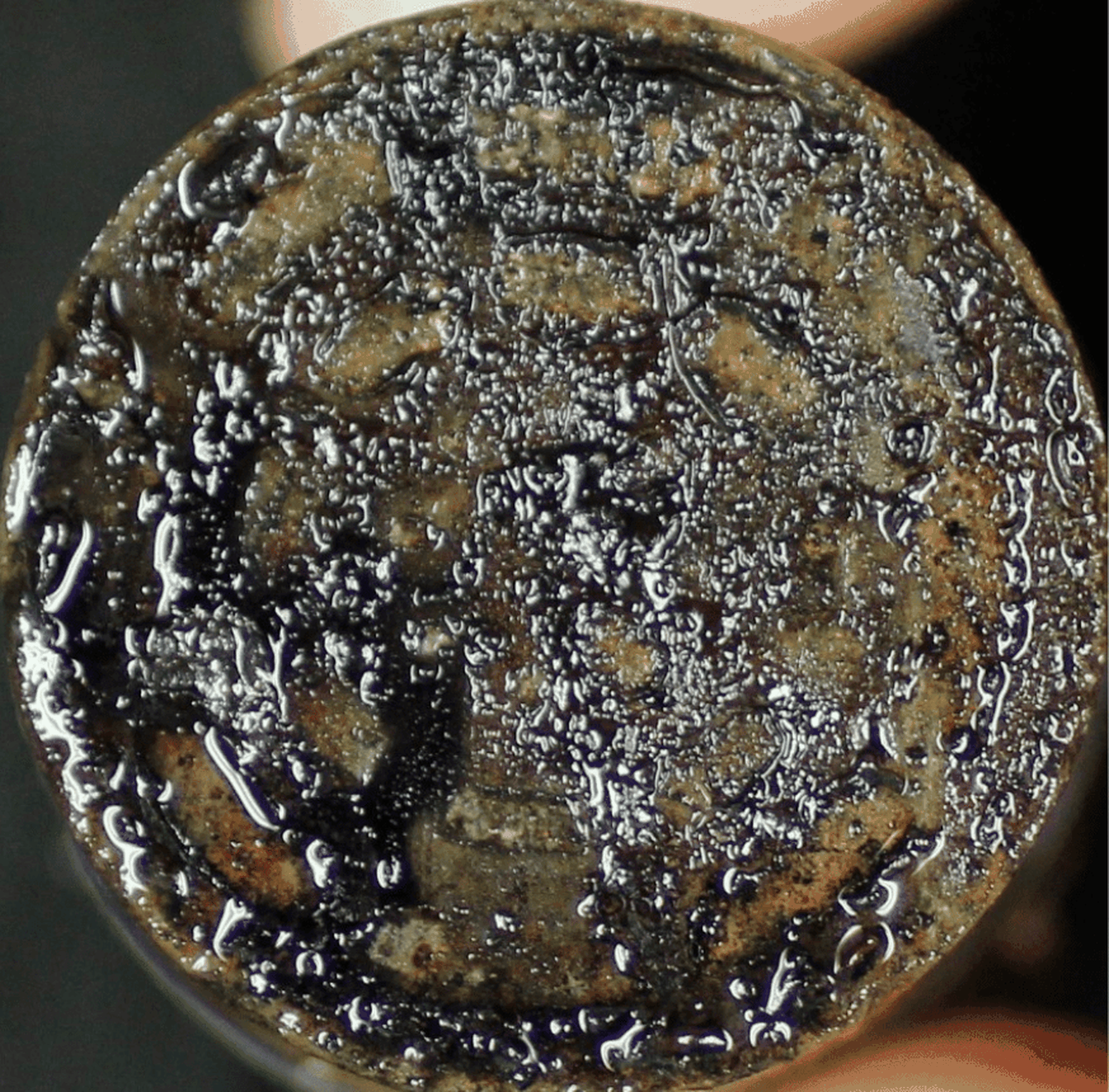 Close-up of a weathered, corroded coin covered in droplets, obscuring most of its details and inscriptions.