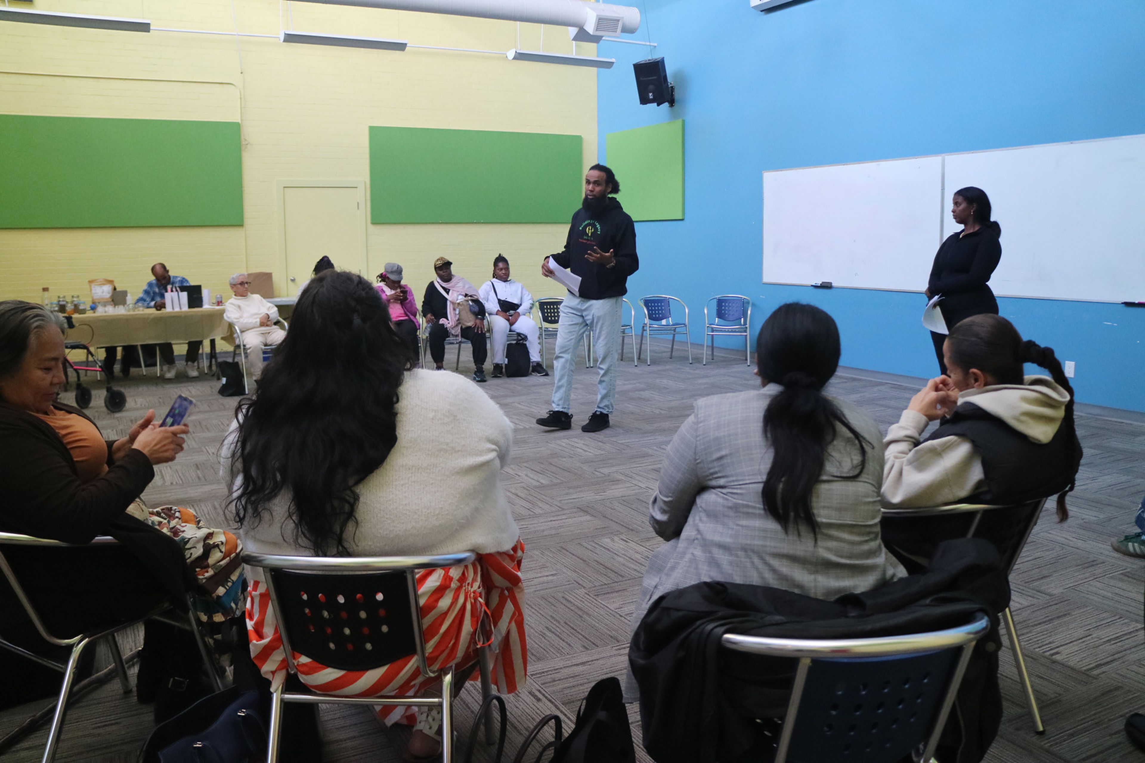 A man stands and speaks to a seated group in a colorful room with green and yellow walls. A woman stands nearby, listening.