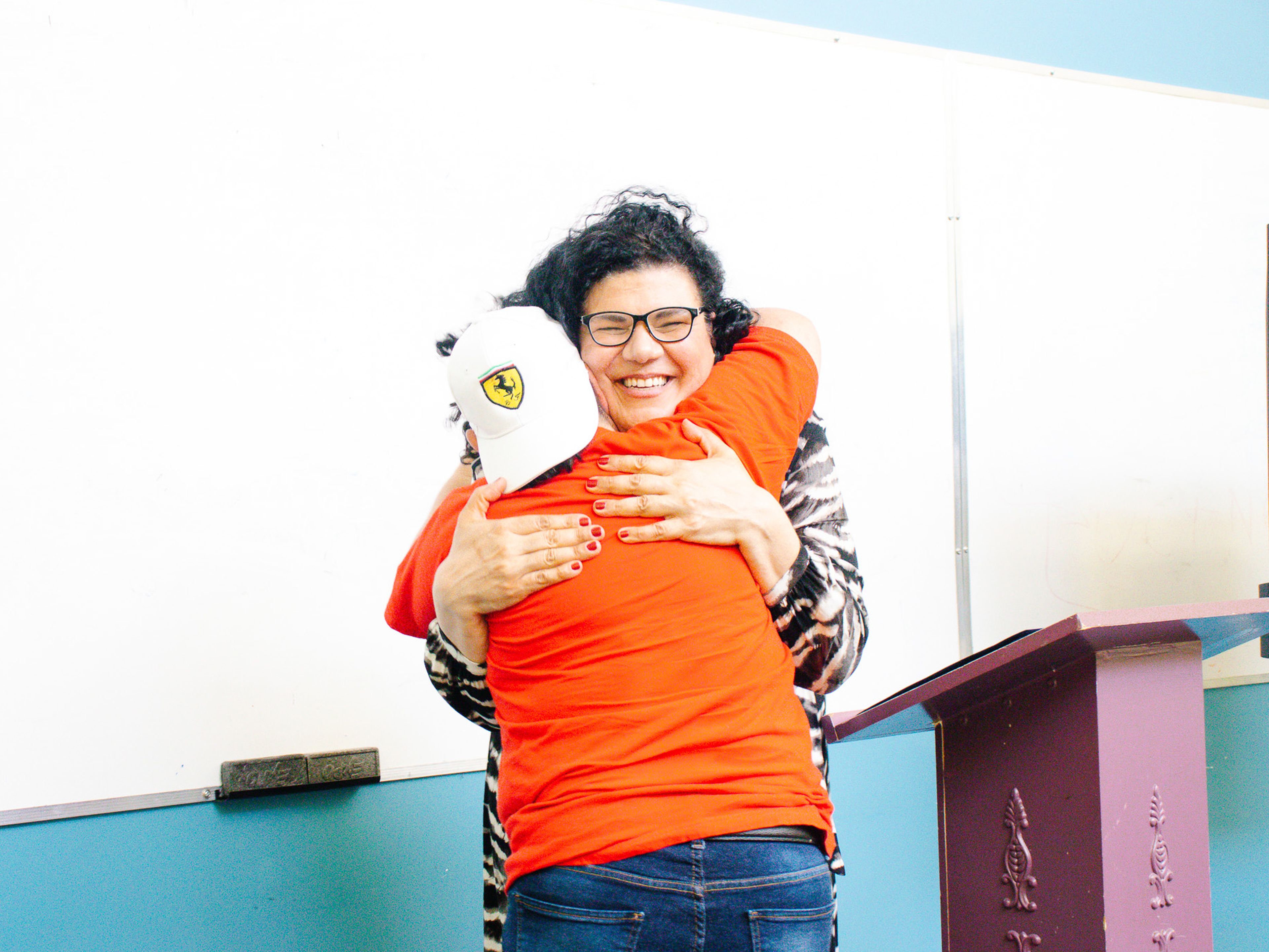 Two people hugging warmly in front of a whiteboard. One wears a red shirt and cap; the other has glasses and a patterned sweater.
