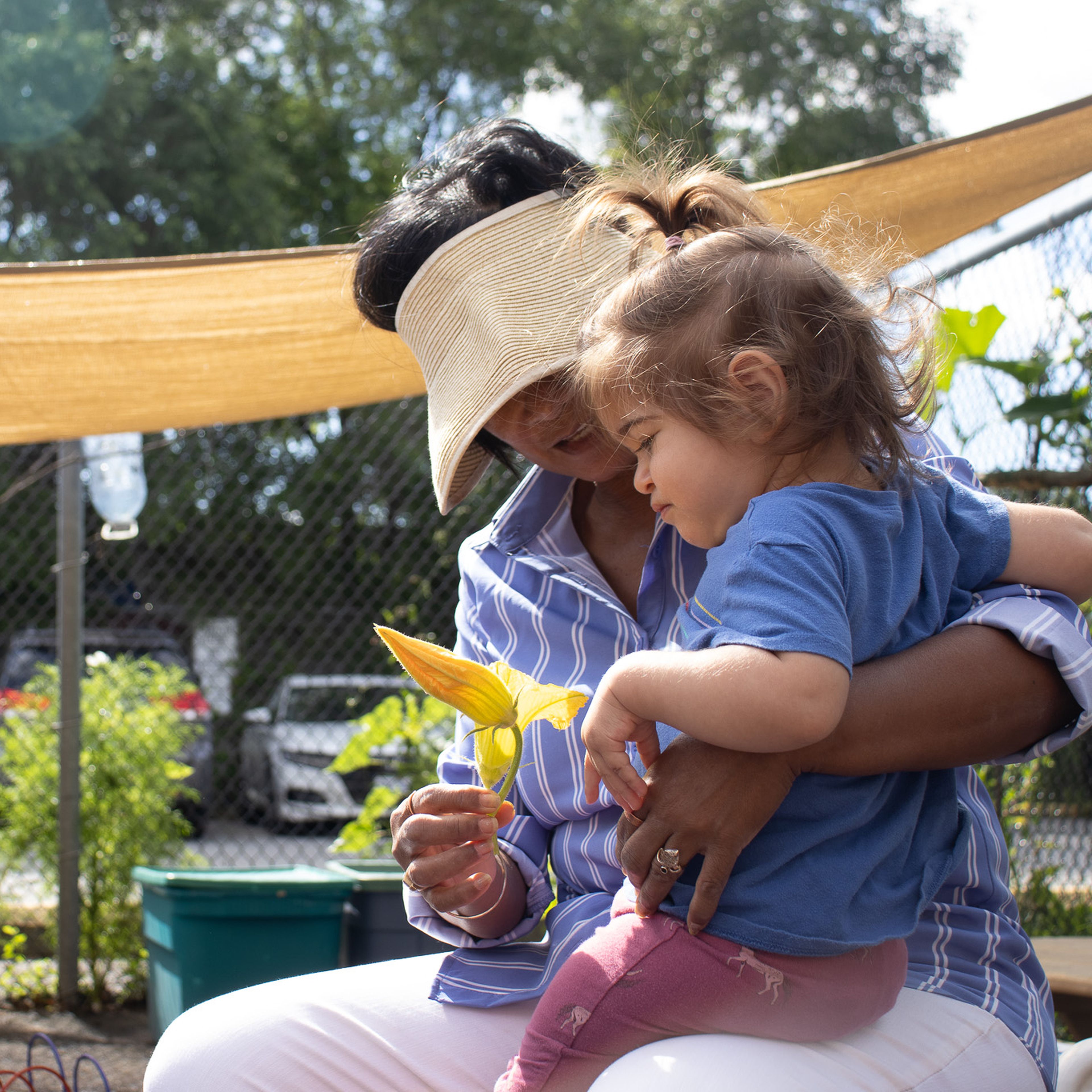 A woman wearing a visor holds a young child in her lap outdoors, both looking at a yellow flower. A playground and trees are in the background.
