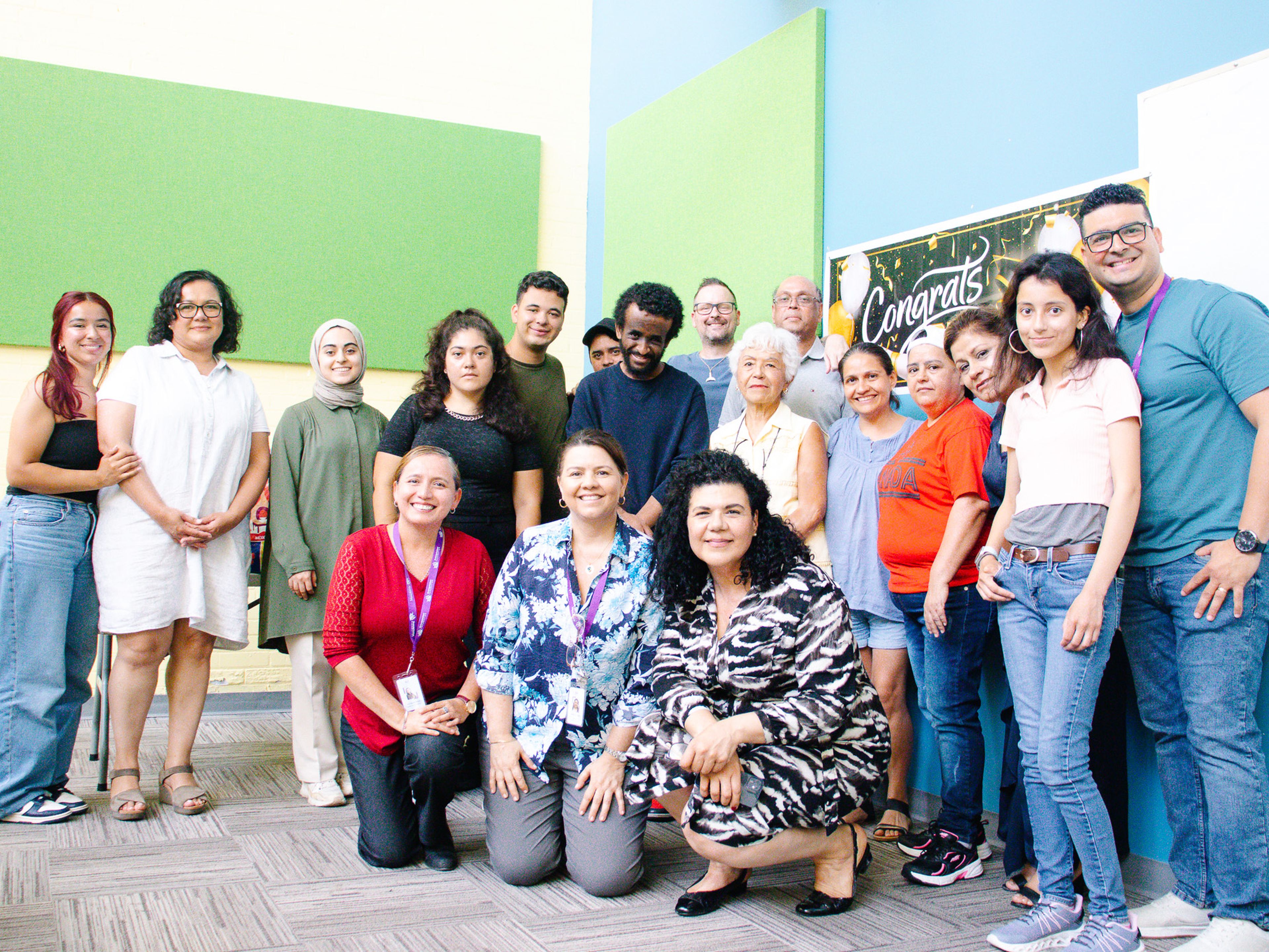 A diverse group of people smiling and posing together in a room with a "Congrats" banner on the wall.