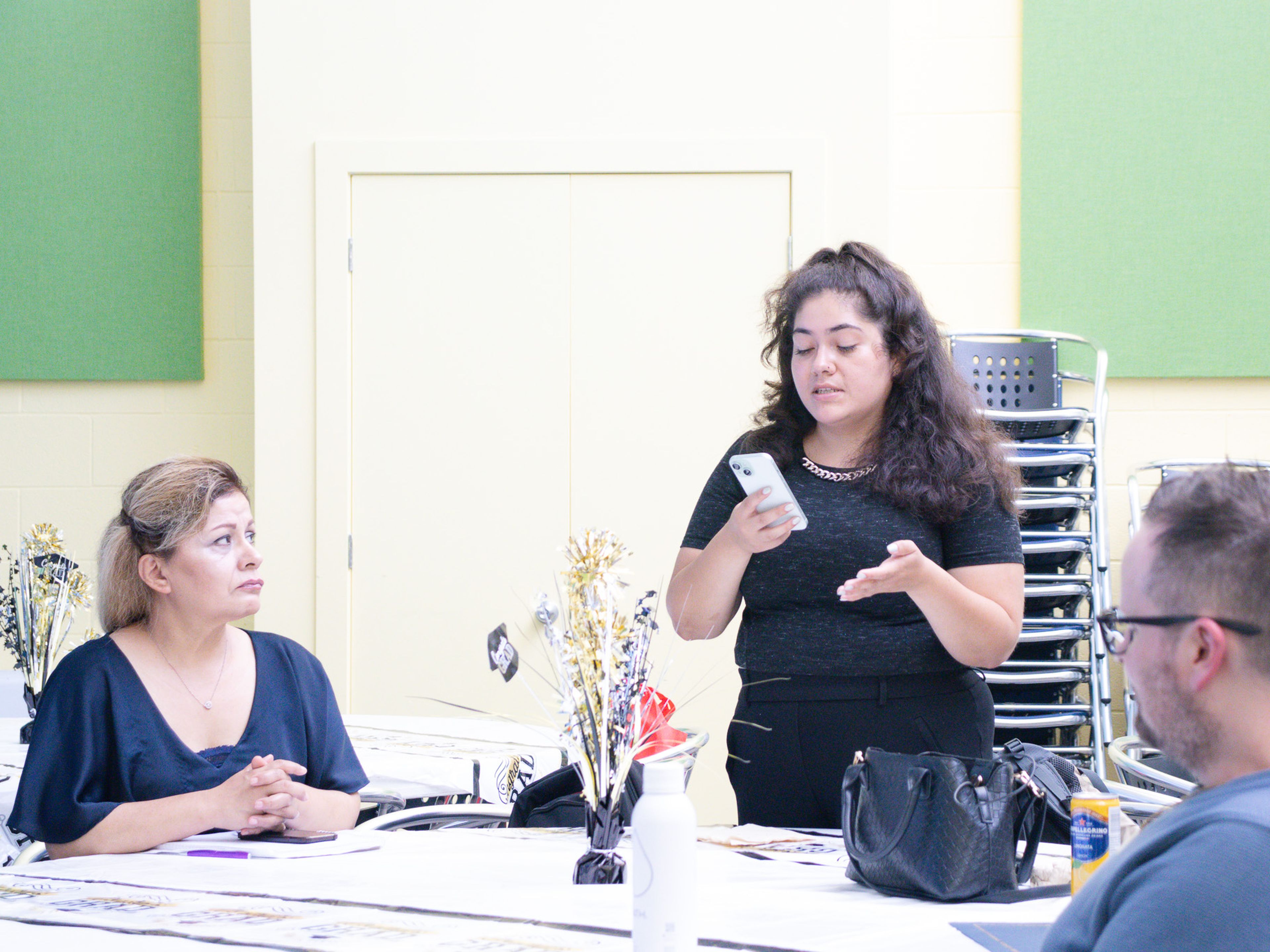 A woman stands speaking, holding a phone, while two people sit at a table listening in a brightly lit room with stacked chairs in the background.