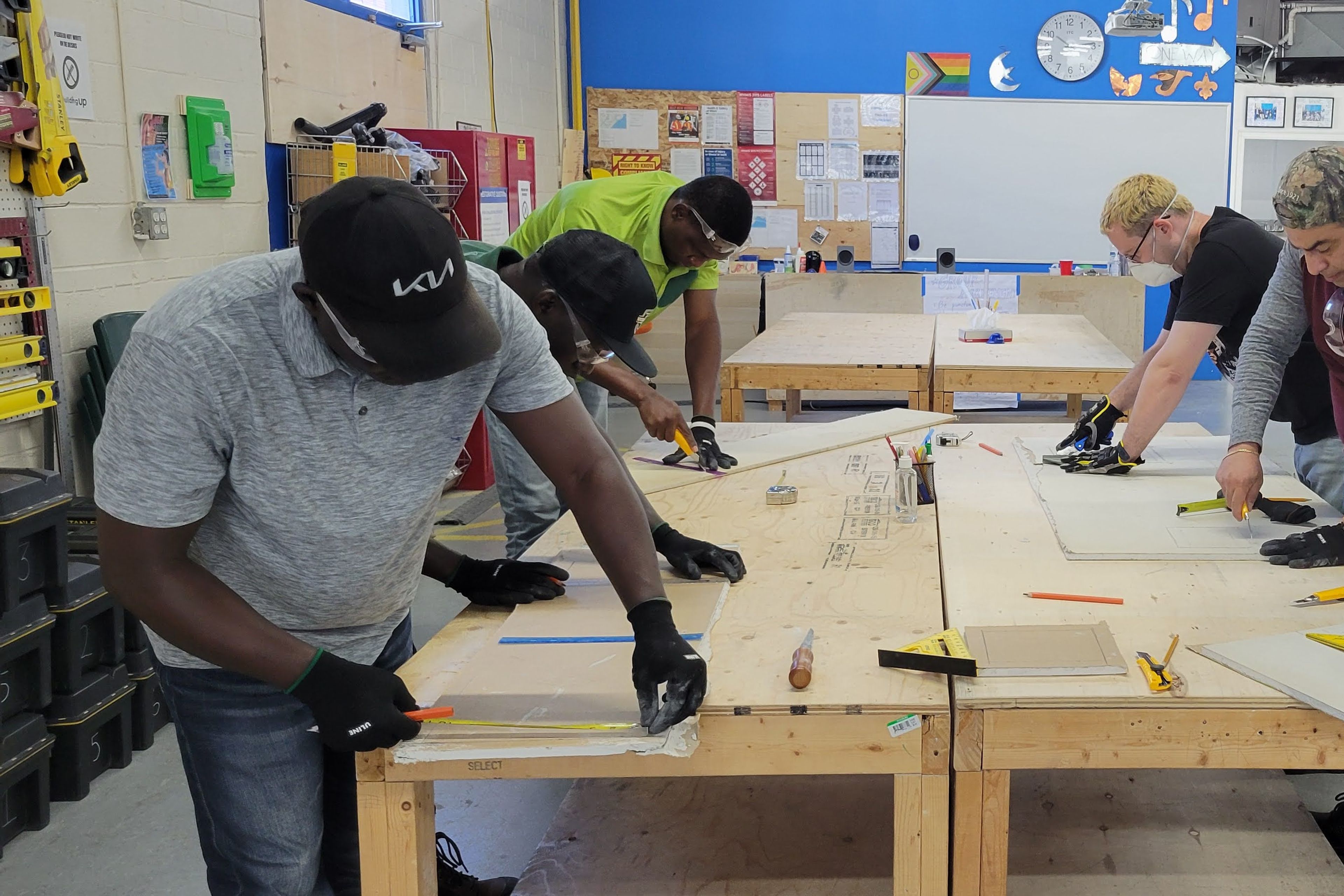 A group of men are standing around a wooden table, all of them are individually working on something, they look like they are making measurements.