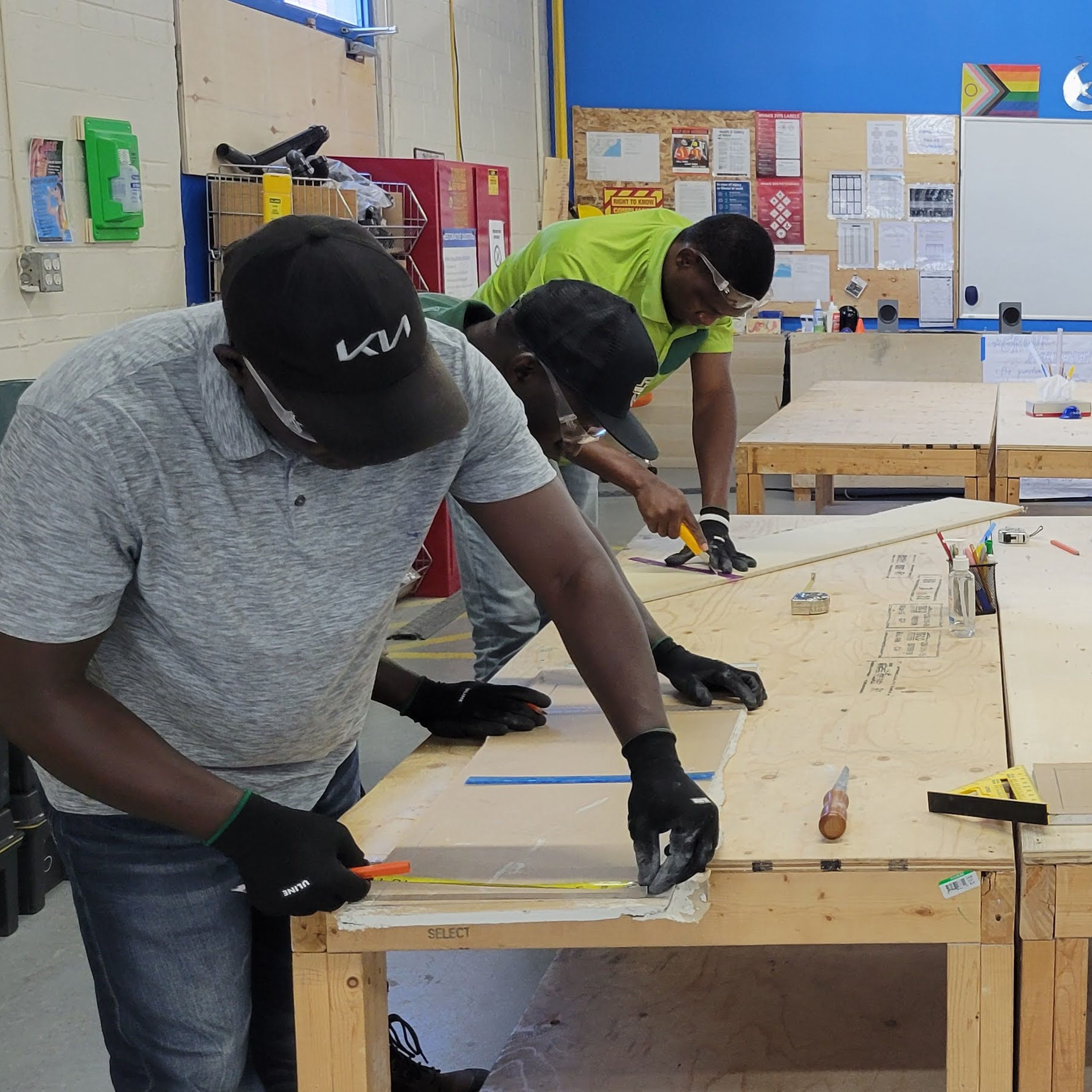 A group of men are standing around a wooden table, all of them are individually working on something, they look like they are making measurements.