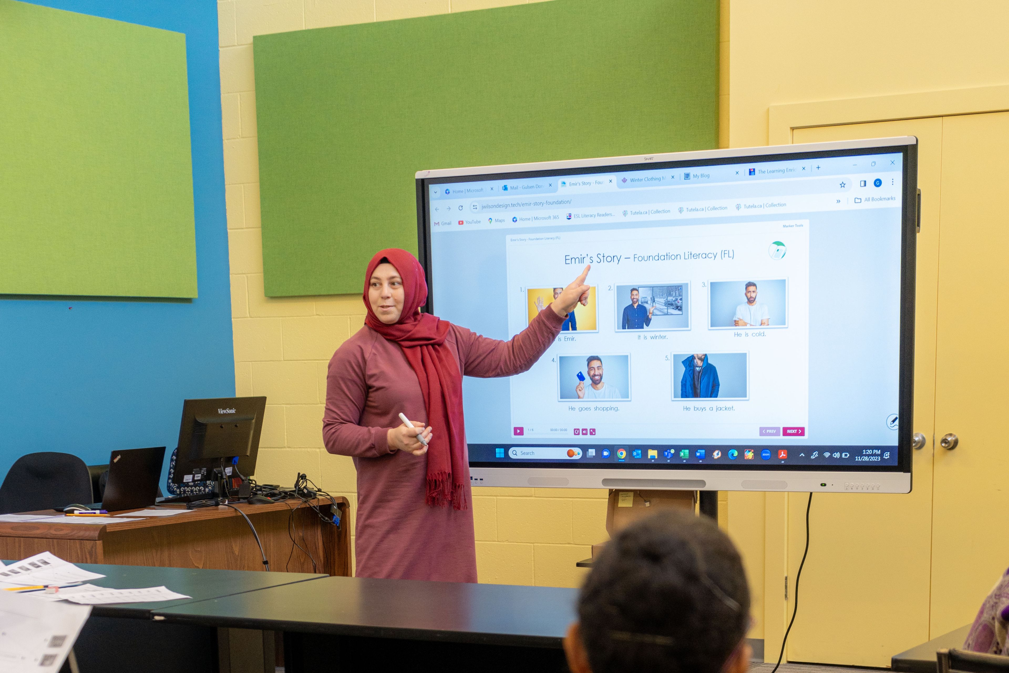 A classroom filled with students sitting at their desks, and the educator is in front pointing at the board.