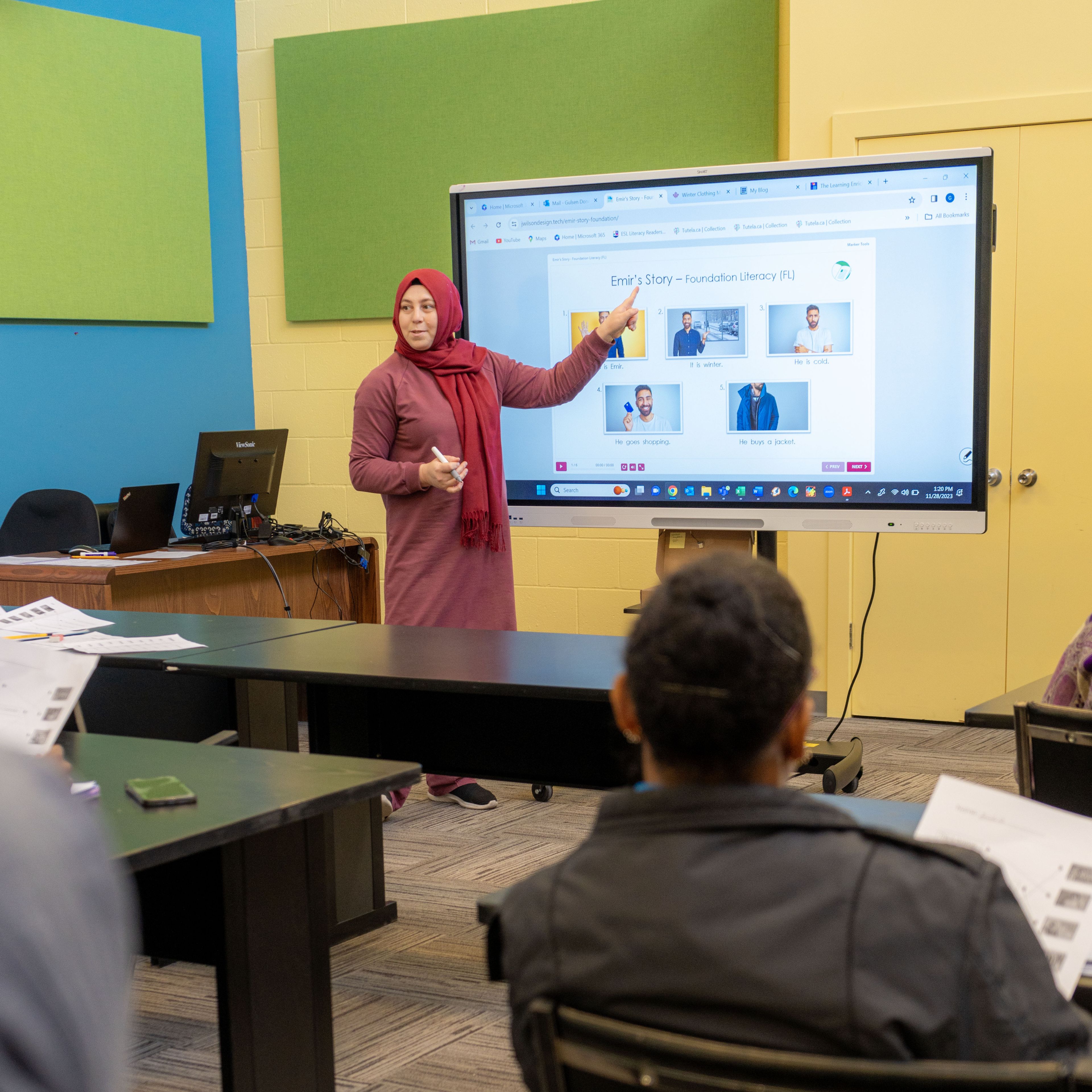 A classroom filled with students sitting at their desks, and the educator is in front pointing at the board.