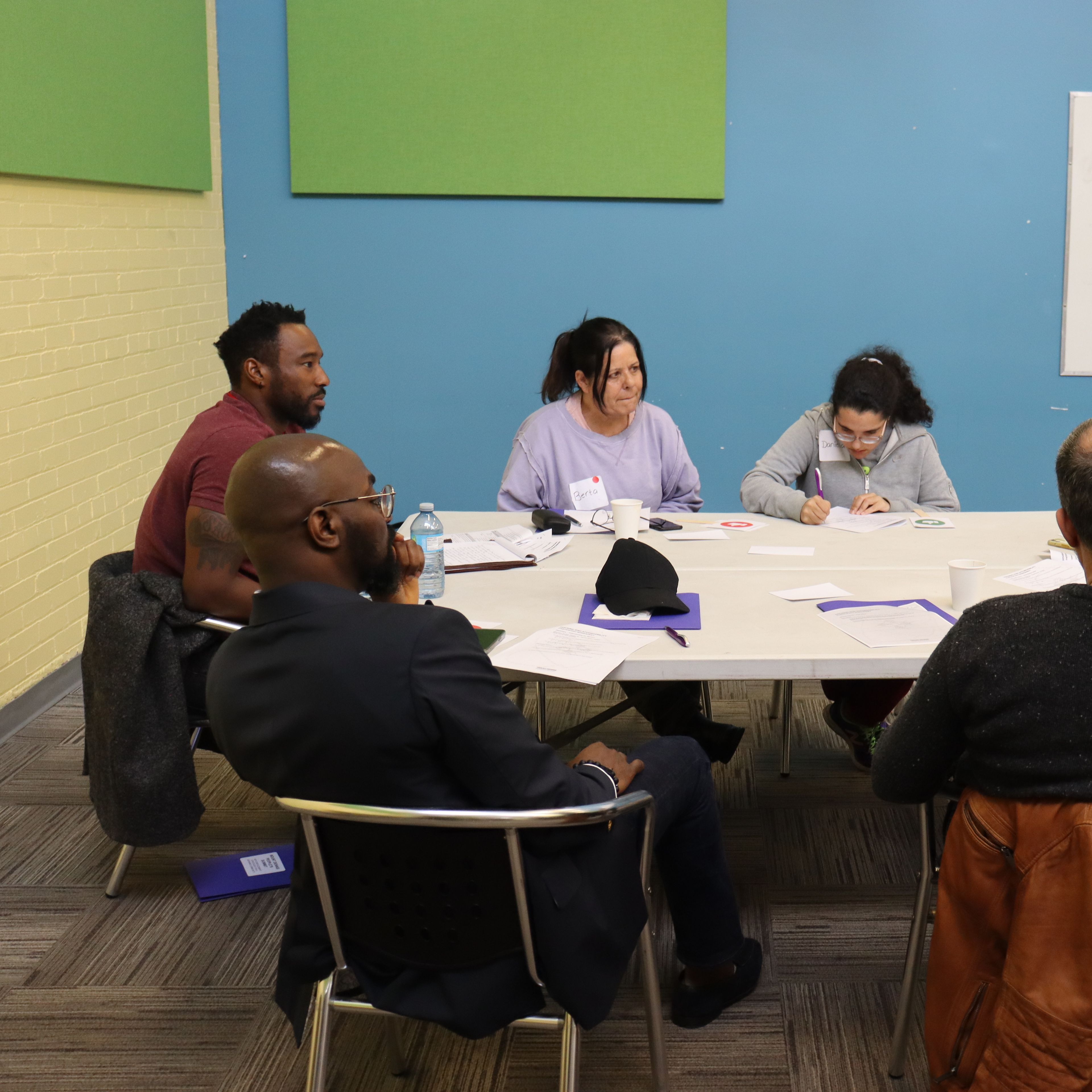 A group of people sitting around a table in a discussion, with notebooks and papers scattered on the table in a brightly colored room.