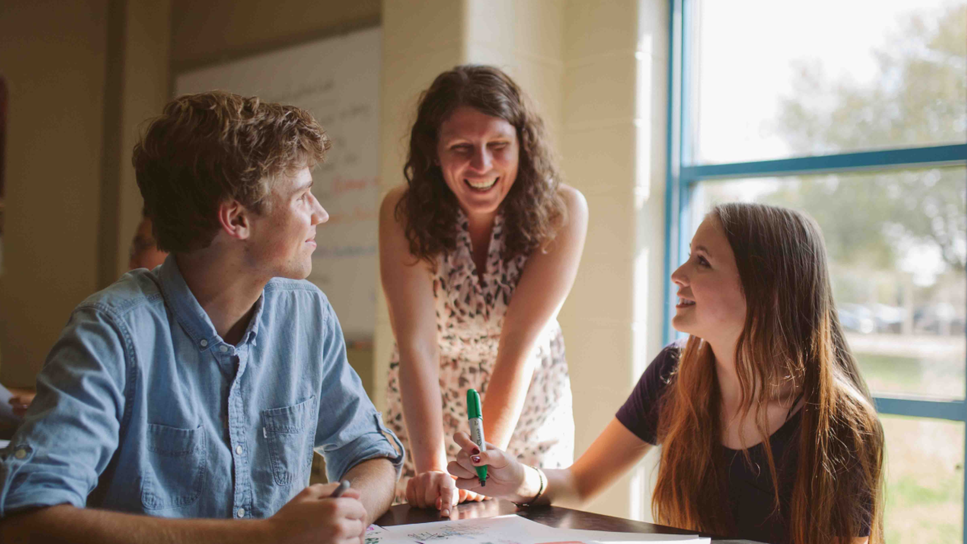 Smiling woman leans over a table helping two students with papers and markers in a sunlit classroom.