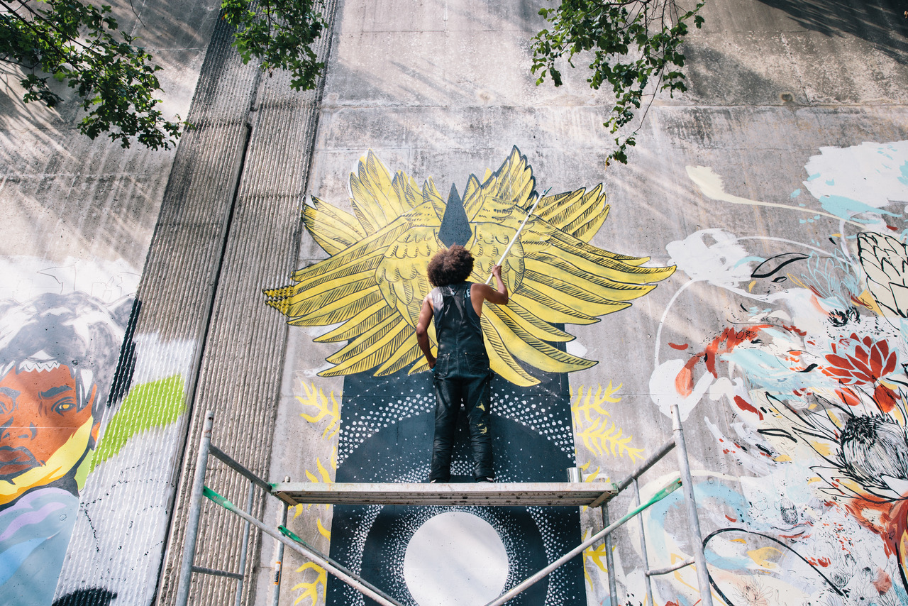 The Foward Warrior Murals along the Southeast Trail are great to stroll along, day or night. (Photo Credit: The Sintoses) Person on scaffolding painting a large yellow winged mural on a concrete wall, flanked by colorful street art.