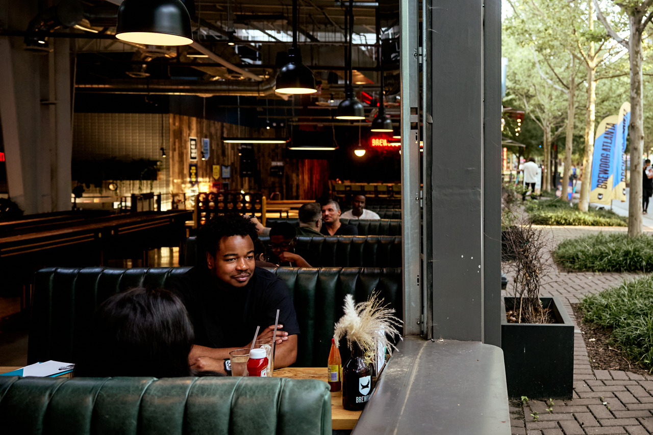People sitting in a cozy restaurant booth near large windows, with drinks and decorative plants on the table, and a view of the Atlanta Beltline.