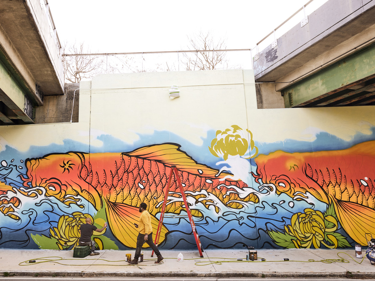 A man sits with his back toward the camera, painting a large colorful mural of fish and water. Another man walks by the artist at work.