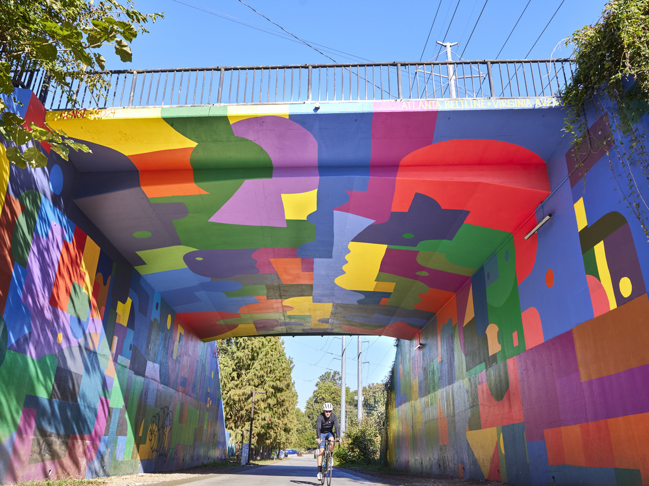 Colorful mural painted on an underpass, depicting abstract shapes and patterns, with a cyclist riding beneath it on a sunny day.