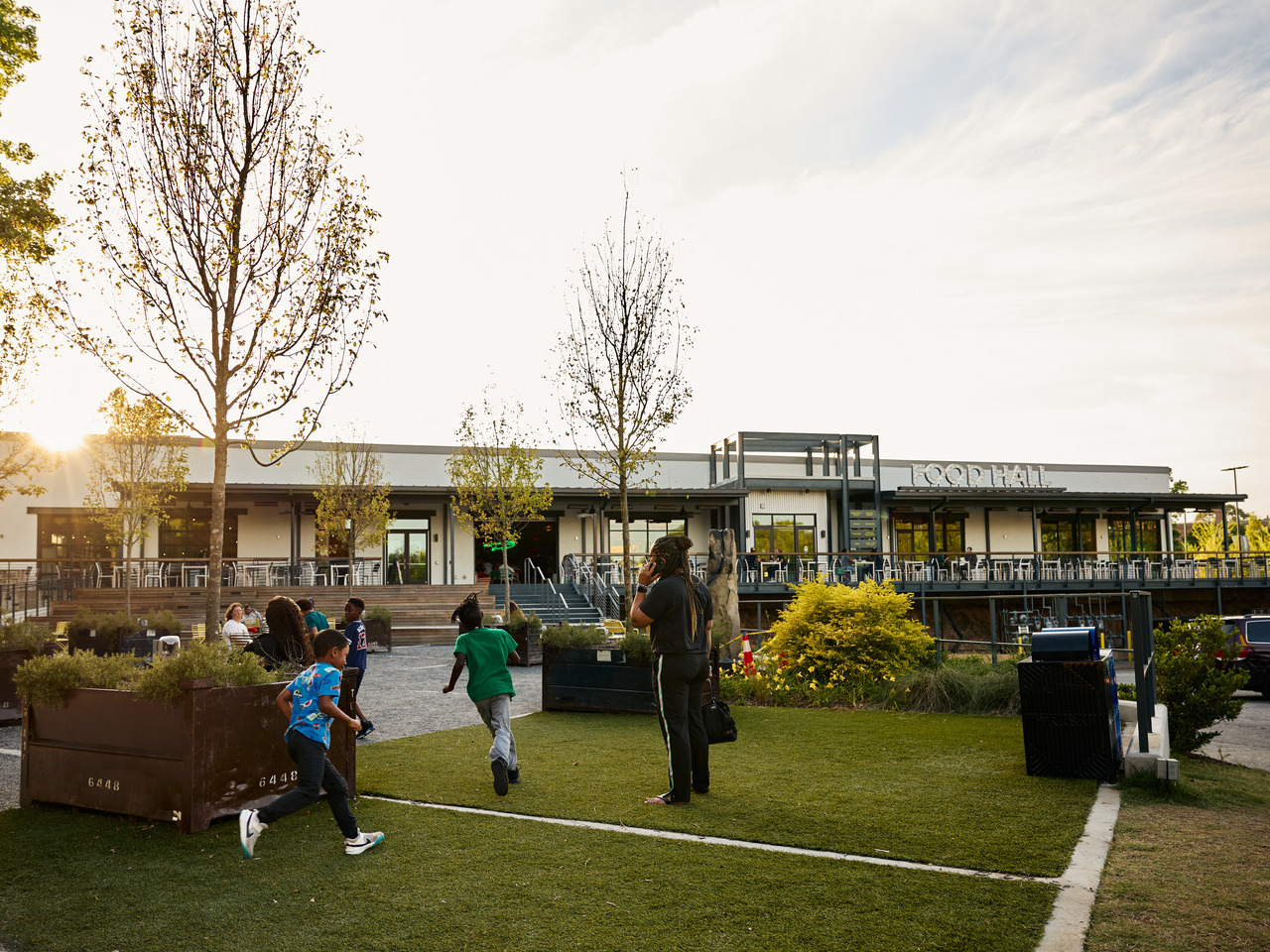 Children playing on a grassy area near a modern food hall with outdoor seating, surrounded by young trees, during sunset.