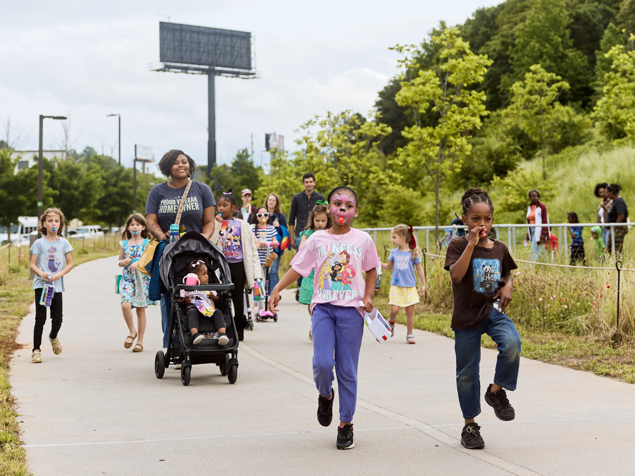 A group of children parade down the Beltline, with one child in a stroller and others holding small items, surrounded by green trees.