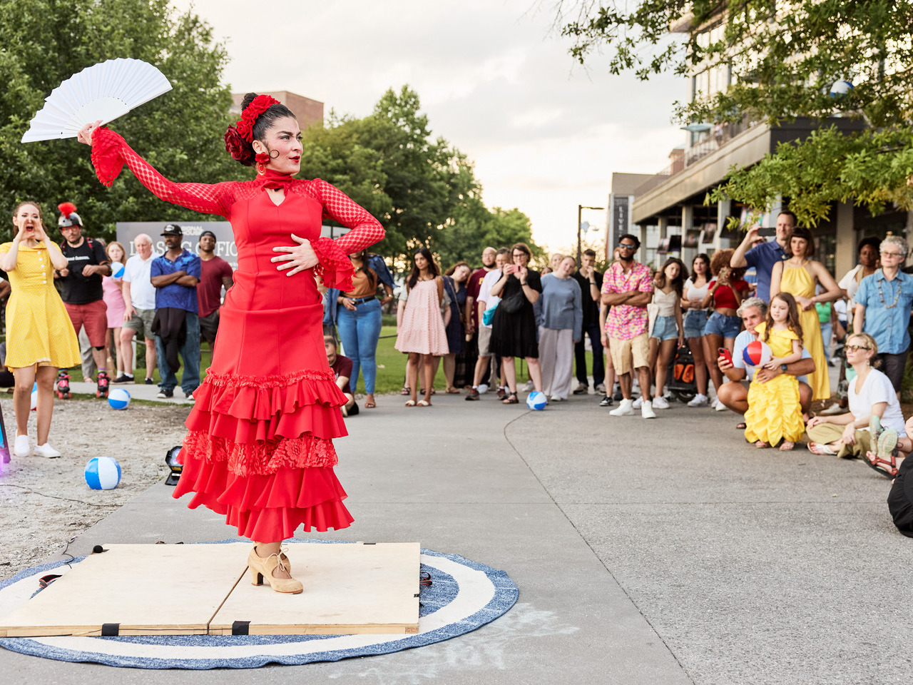 A performer in a red dress dances with a fan on the side of the Eastside Trail, surrounded by an audience under a cloudy sky.