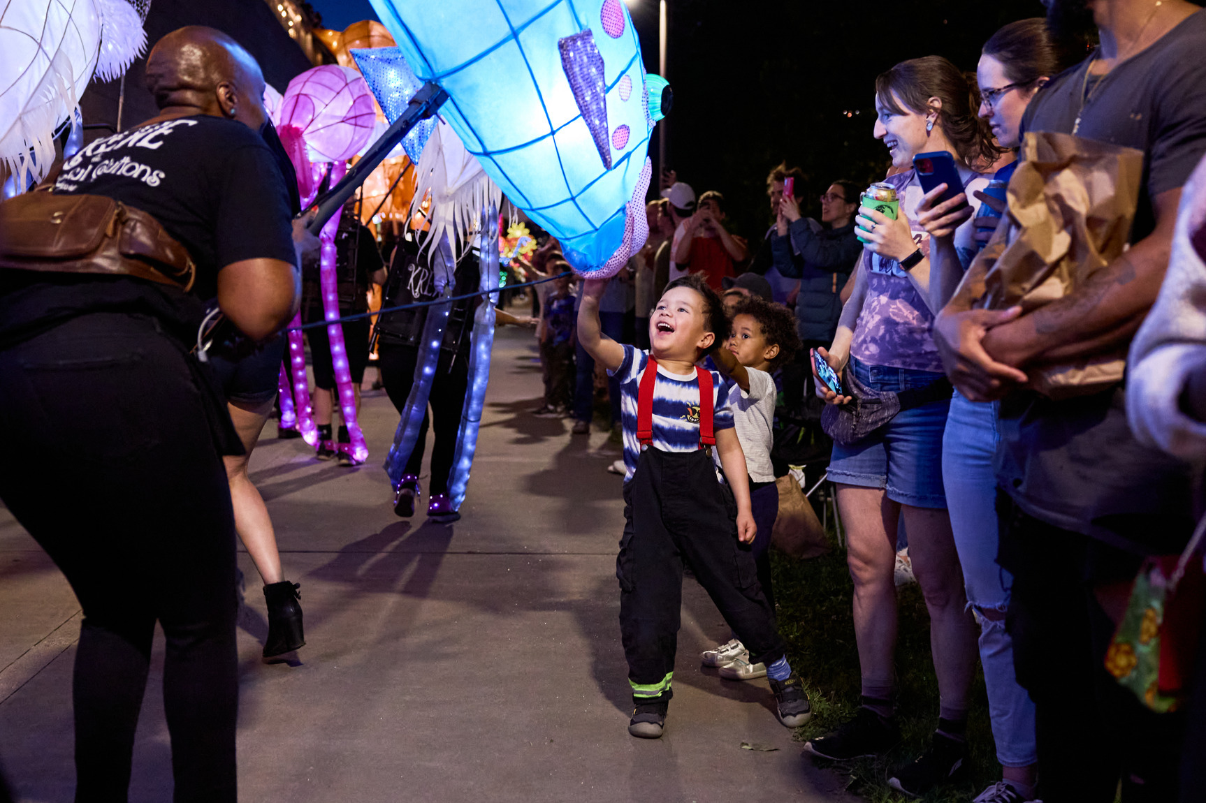 A child excitedly reaches for a glowing lantern held by a performer during a lively parade, with a crowd watching and smiling.