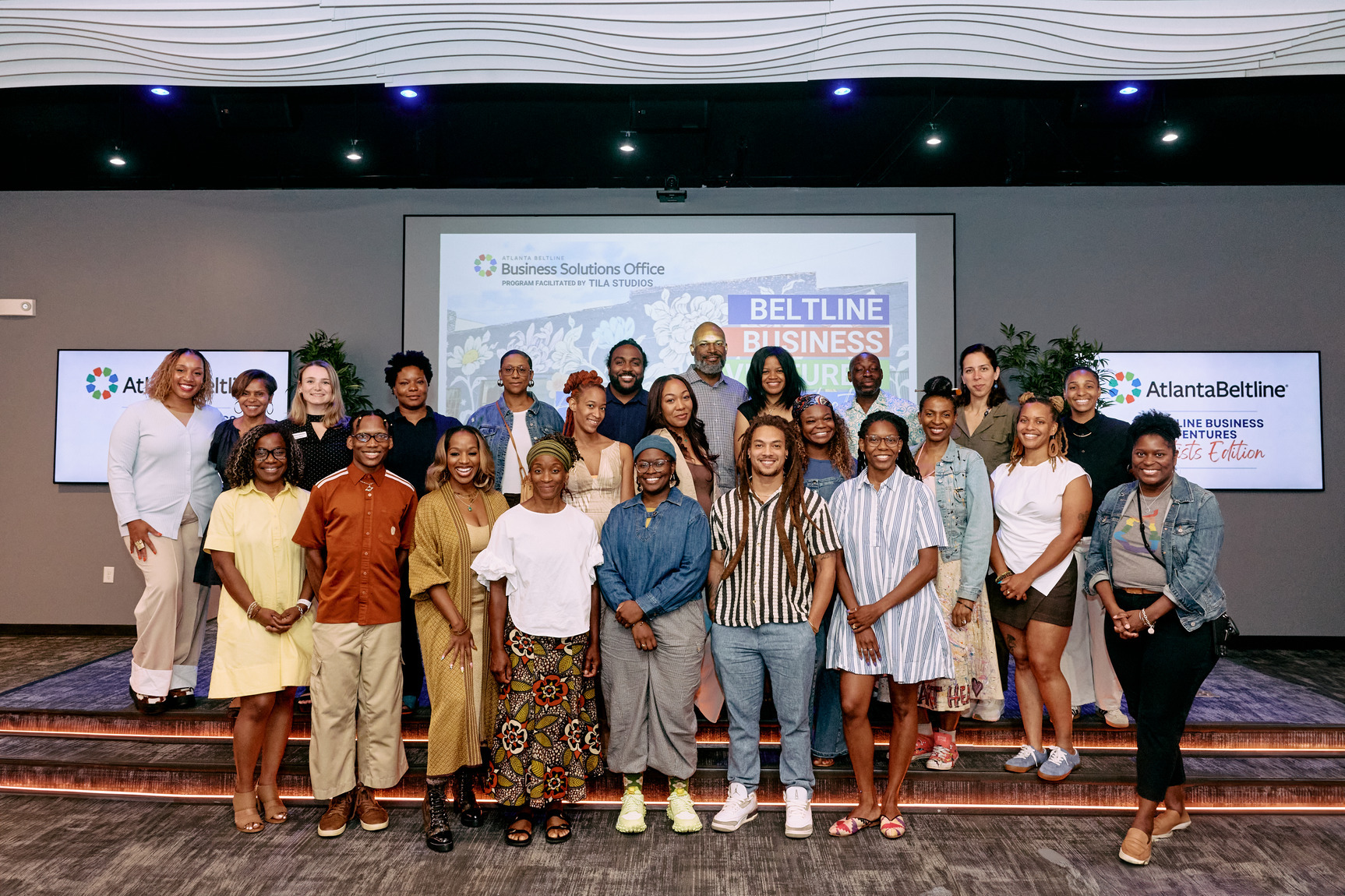A diverse group of people smile and pose in front of a presentation screen displaying "Beltline Business Ventures" at an indoor event space.
