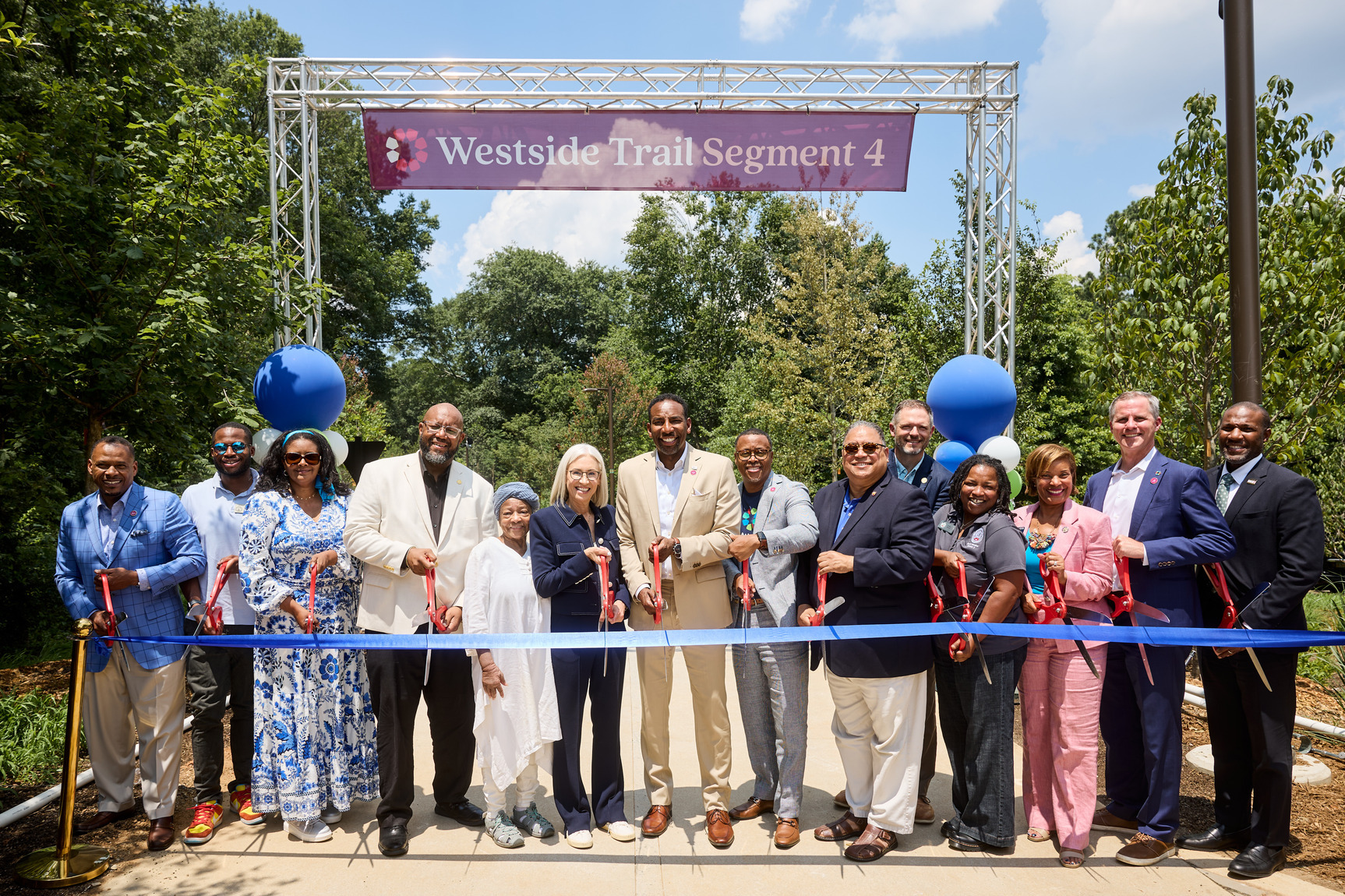 A photo of a group of people posing together, holding giant scissors, and standing behind a big blue ribbon. Behind them is Westside Trail - Segment 4.